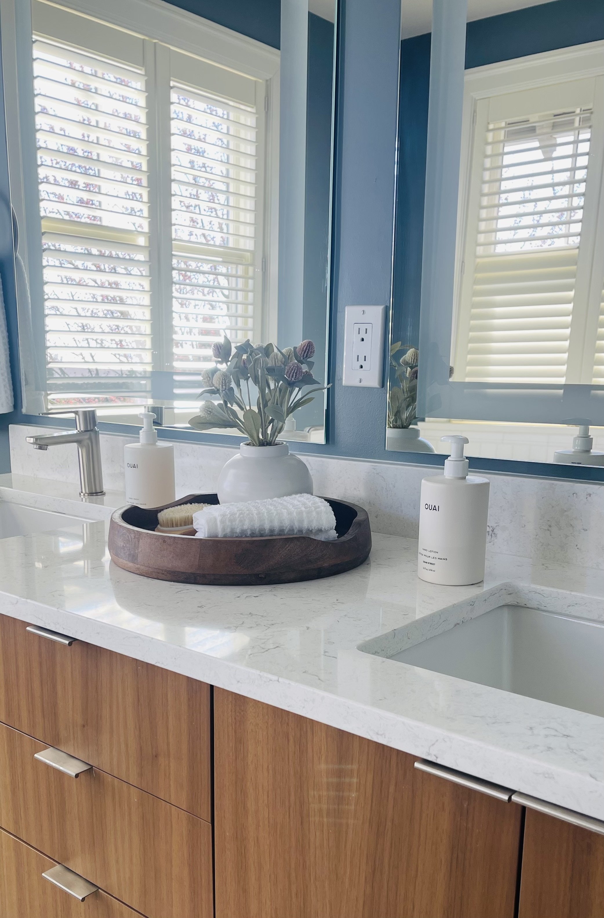 Bathroom countertop with a white soap dispenser, a white lotion bottle labeled 'OUAI', a white towel, a small brush, a potted plant, and a large mirror reflecting windows with white shutters and a blue wall.