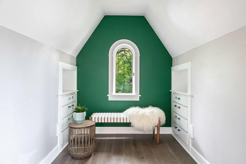 Small attic room with a green accent wall, white built-in shelves, and a window with a decorative black grille. The room has a side table with a potted plant, a bench, and a fluffy white throw on a small wooden stool.