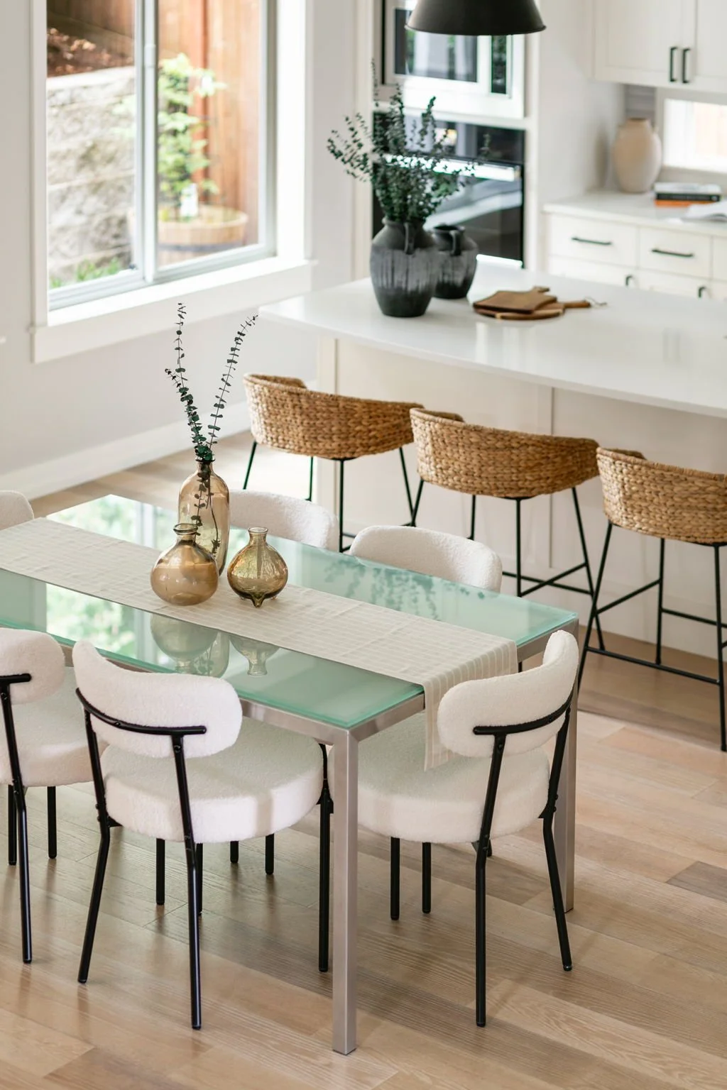 Modern dining area with a glass-top table surrounded by white cushioned chairs and decorative vases, overlooking a bright kitchen with barstools and a large window.