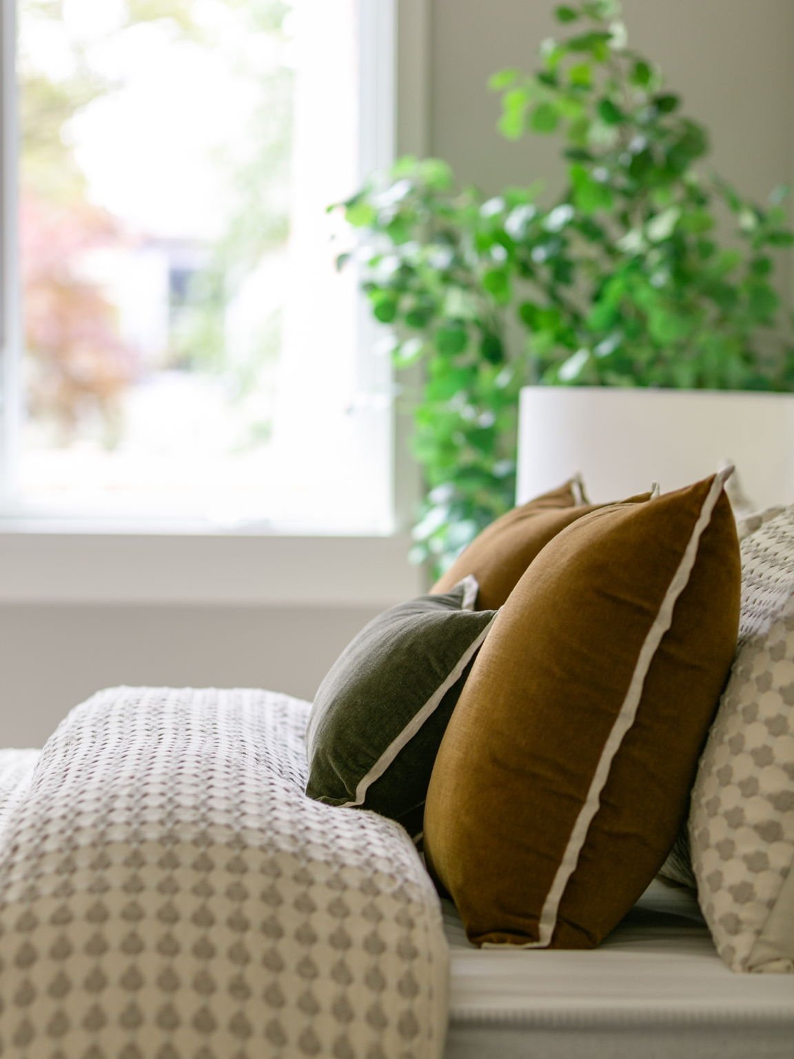 A cozy bedroom with a white bed frame, beige and brown pillows, a patterned blanket, and a green plant near a window with natural light.