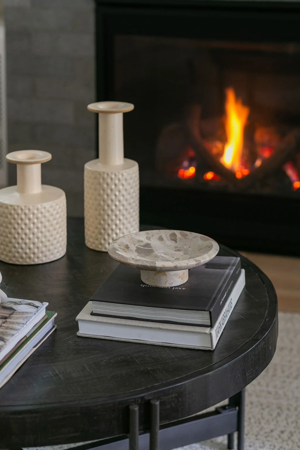 A black round coffee table with books, a marble cake stand, and decorative vases on it in front of a lit fireplace.