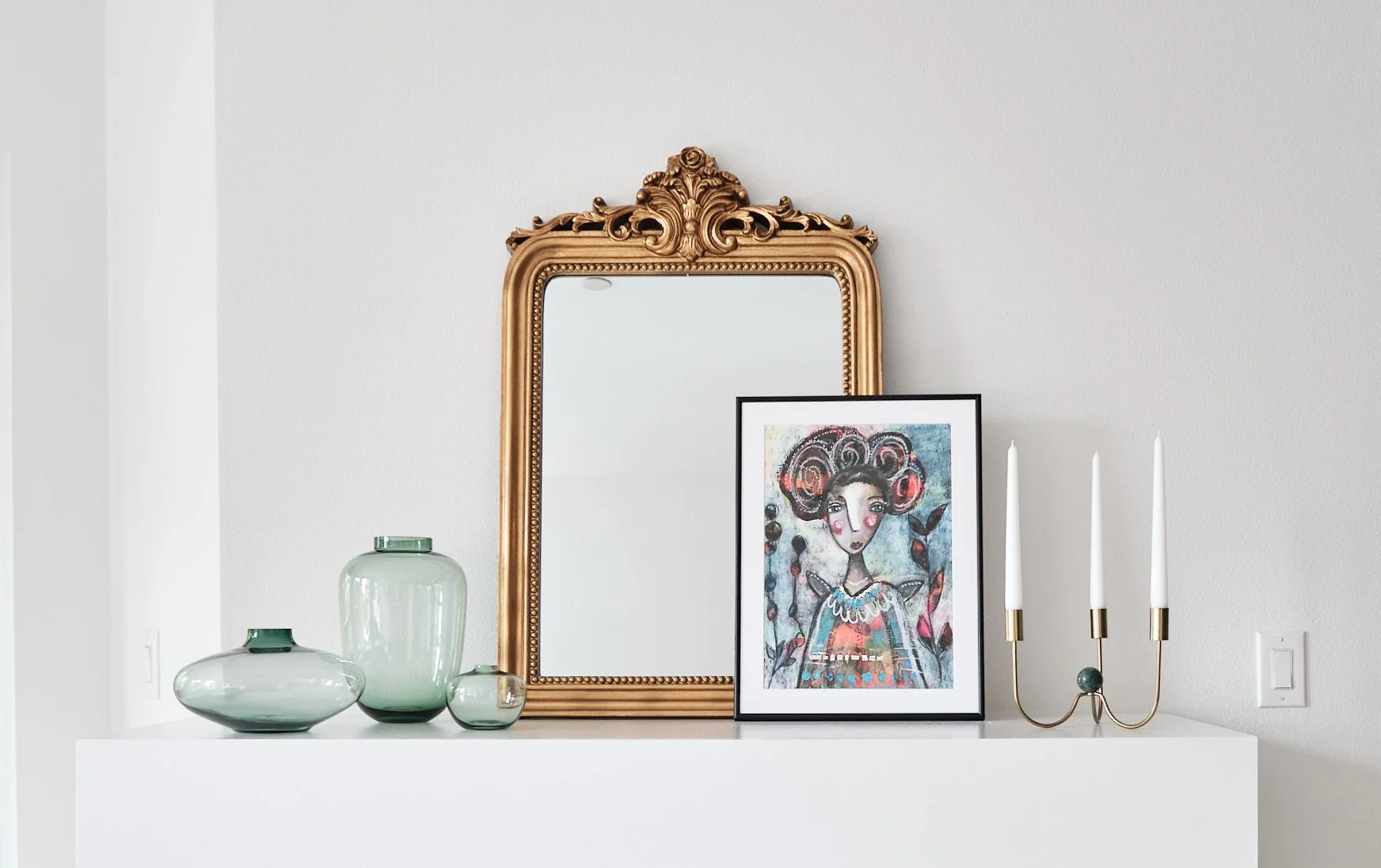 Decorative wall arrangement featuring a large ornate gold mirror, a framed colorful artwork of a woman with large hair, three white candles in a gold and green candle holder, and four green glass vases of different sizes on a white shelf against a pl