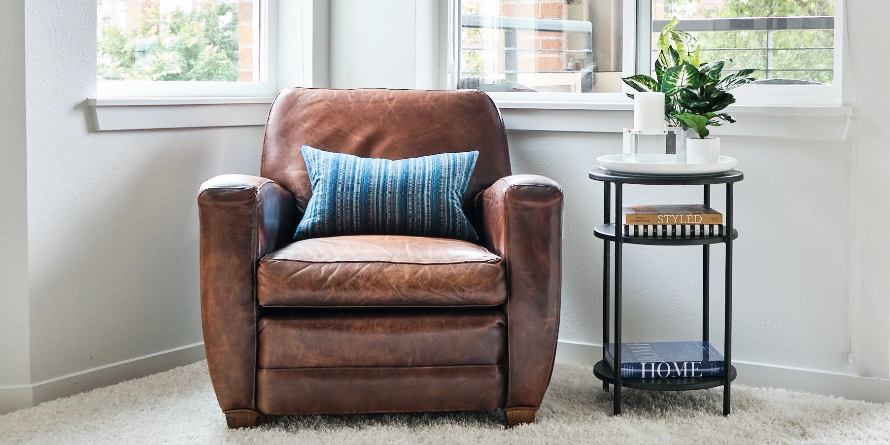 A cozy living room corner with a brown leather armchair and a side table. The armchair has a blue striped pillow. The side table has a white tray with two white candles and a green houseplant. Behind, there are large windows with a view of a balcony 