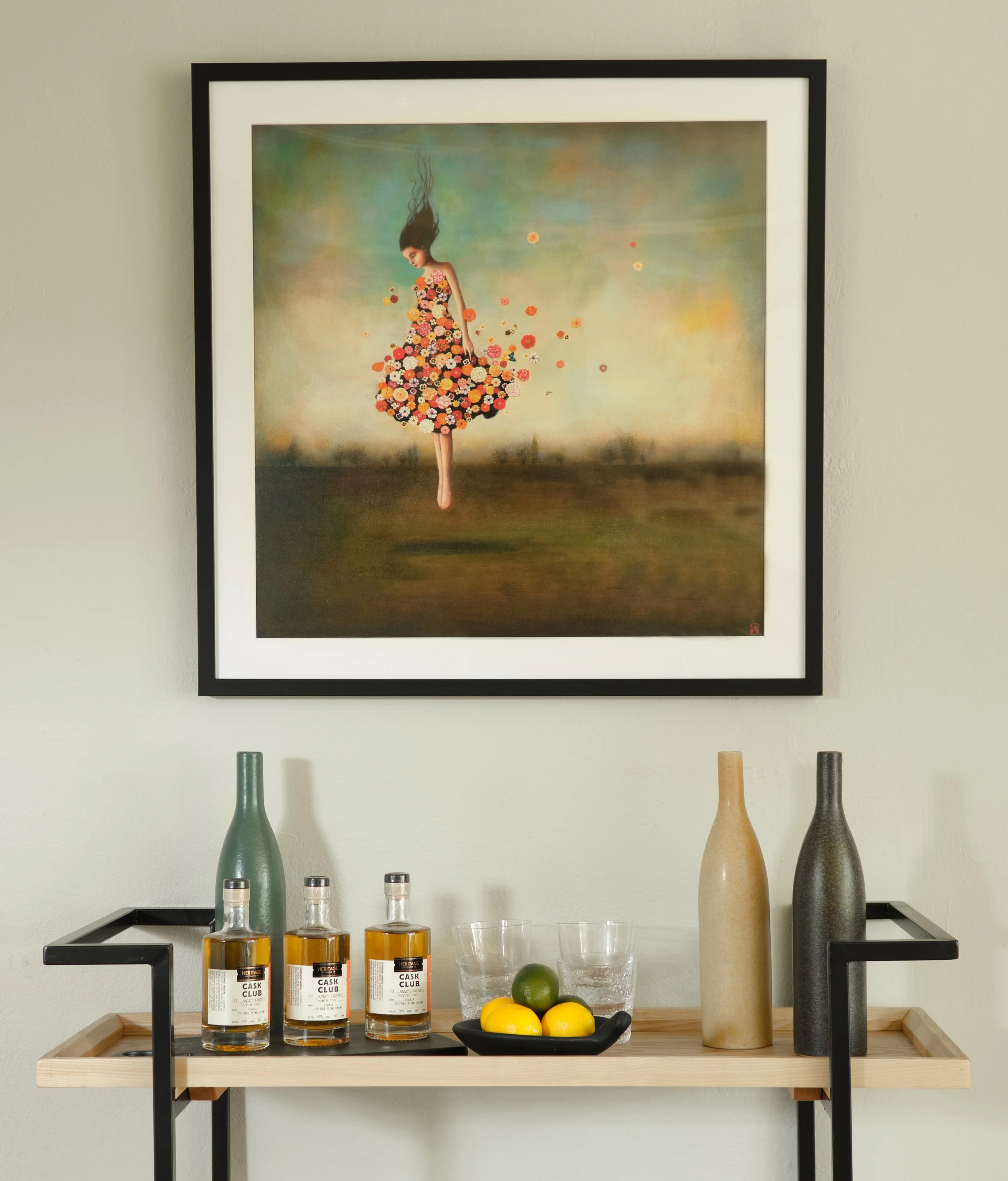 A wooden table with a black metal frame holding three bottles of whiskey, a lime, two lemons, a clear glass bowl with lemons, a clear glass with water, and two empty glasses. Behind the table is a painting of a woman wearing a dress made of flowers, 