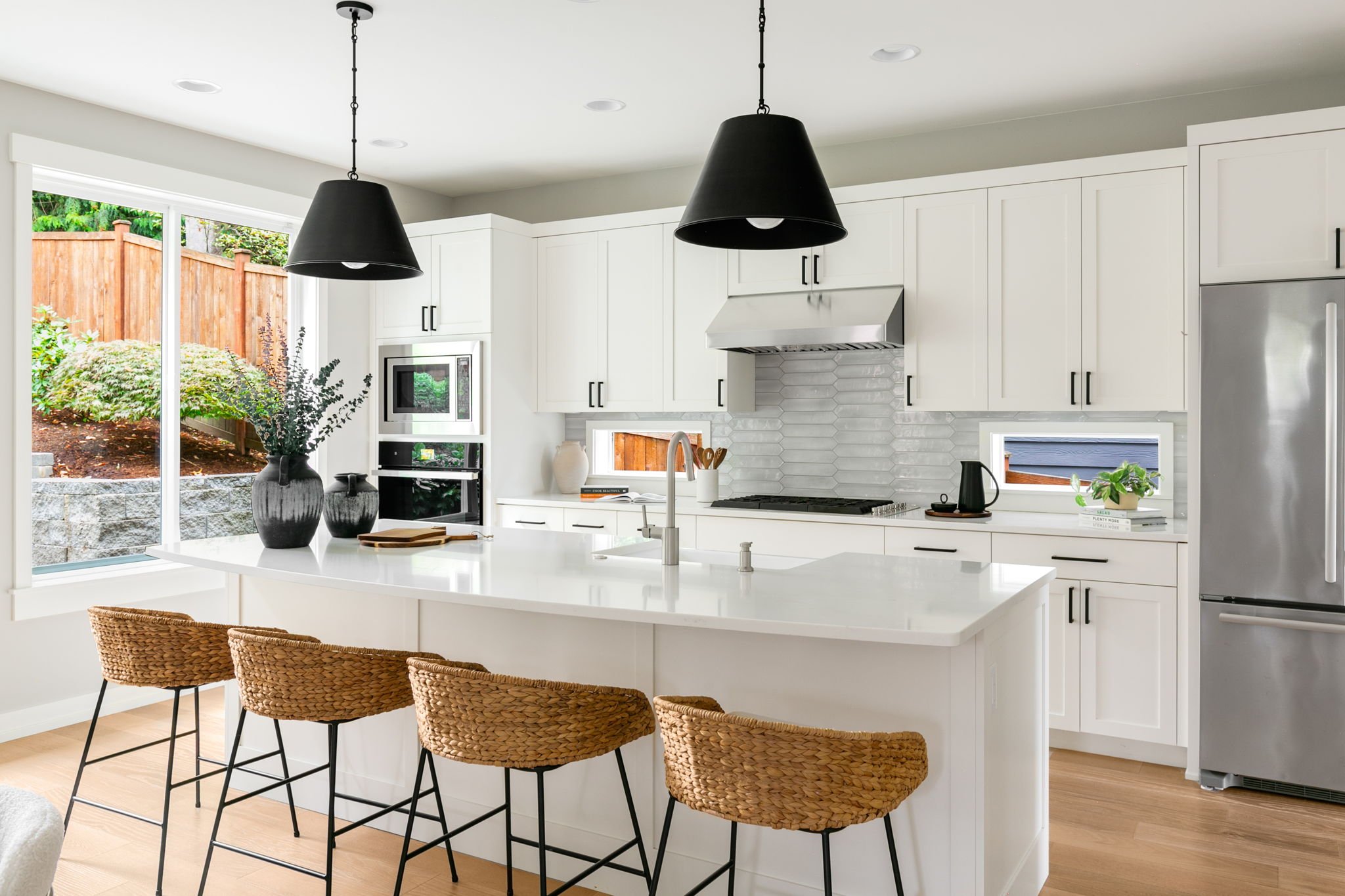 Modern kitchen with white cabinets, black hardware, large kitchen island, black pendant lights, stainless steel refrigerator, microwave, and oven, with a window overlooking a backyard with a wooden fence and greenery, and woven bar stools at the isla