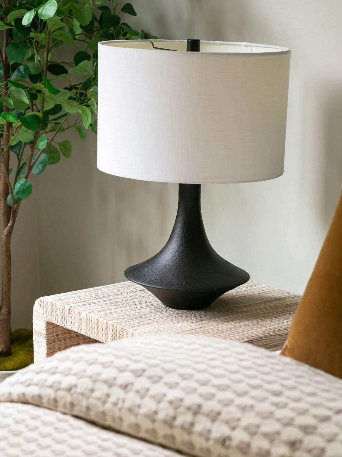 A black ceramic table lamp with a white fabric lampshade sitting on a beige textured side table in a living room, with a green plant in the background.