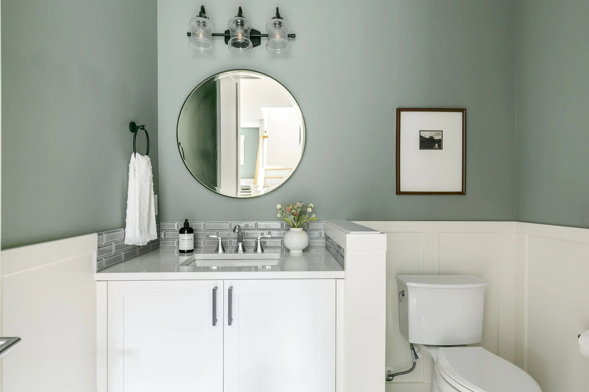 A modern bathroom with a white vanity, grey subway tile backsplash, oval mirror, and a wall-mounted light fixture with three bulbs in glass holders. There is a white towel hanging on a black ring on the wall, a black soap dispenser on the vanity, a w