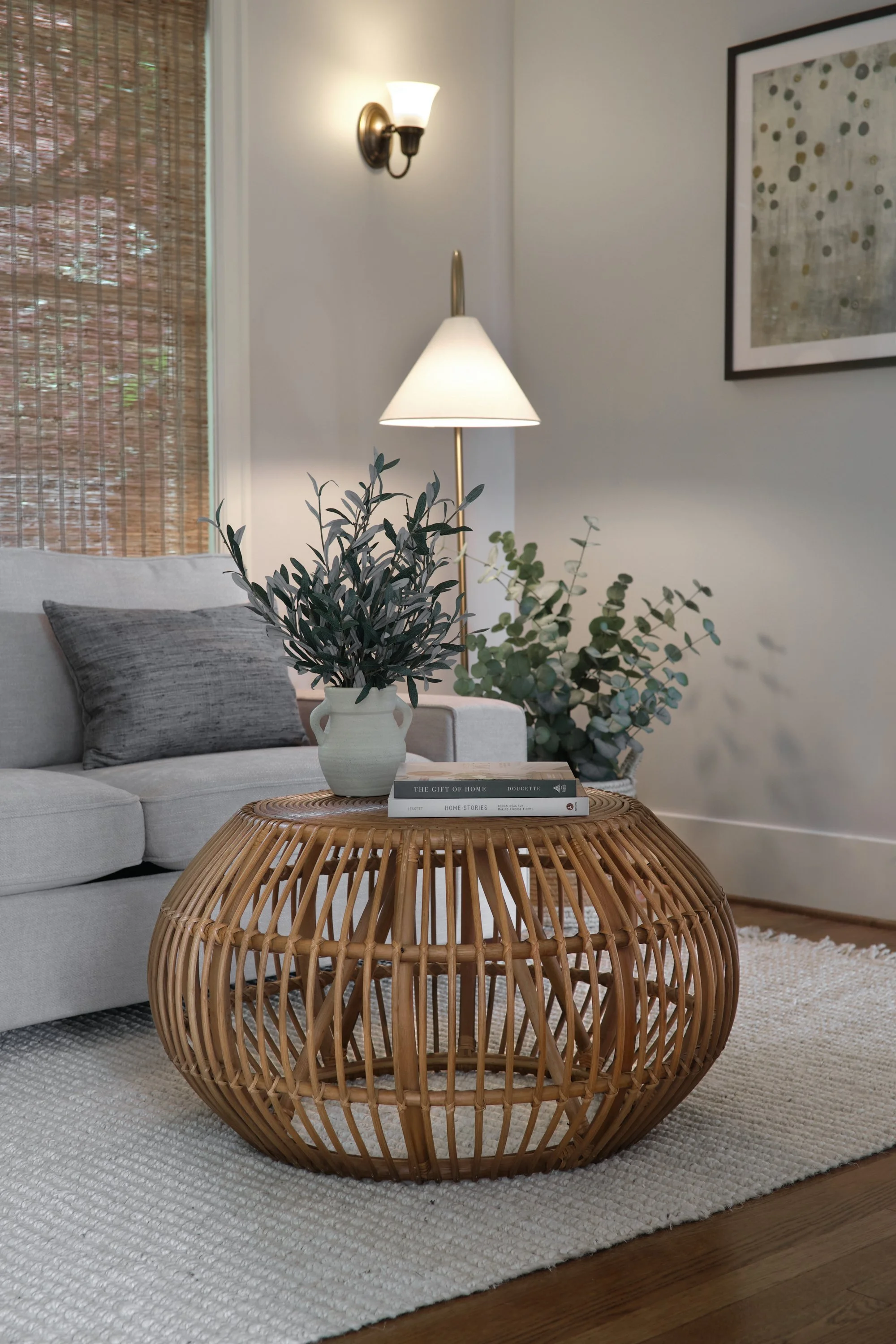 A living room with a white sofa, gray pillows, a wicker coffee table, a potted plant, a lamp, and framed wall art. There is a window with bamboo blinds and a rug on a wooden floor.
