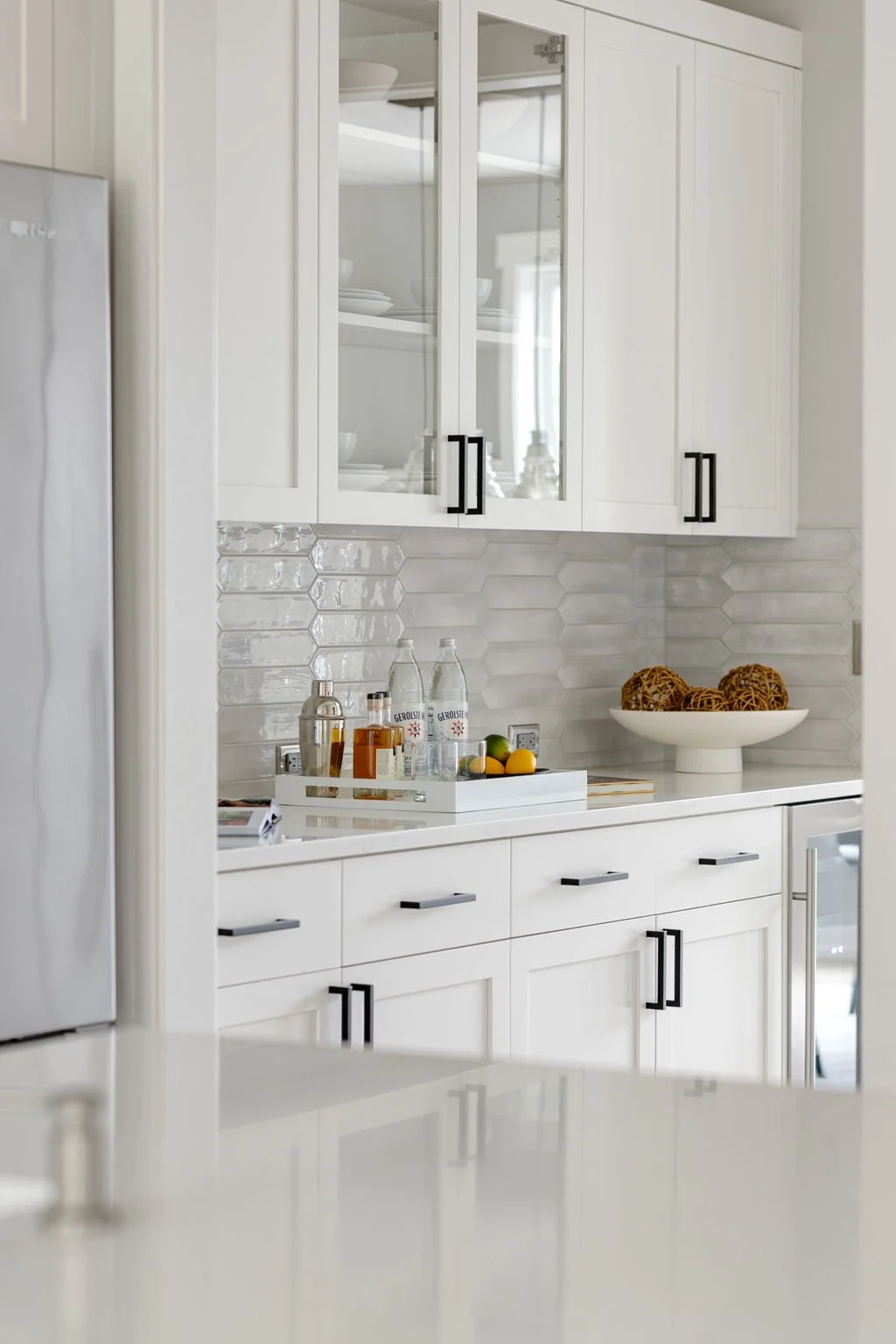 Modern kitchen with white cabinets, hexagonal tile backsplash, and a white countertop featuring a tray with bottled beverages, fruits, and decorative items, as well as a bowl of decorative balls.