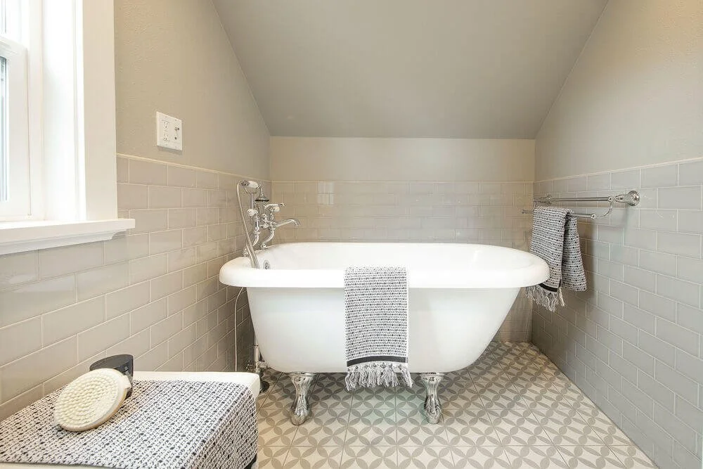 A bathroom with a vintage white bathtub on clawfoot legs, gray tiled walls, and geometric patterned floor tiles. There are gray towels hanging on a rod, a woven basket with a gray towel, and a small window on the left.
