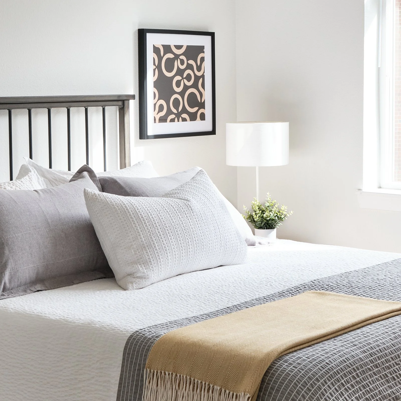 Bedroom with a bed, gray pillows, white bedding with a black-and-white patterned throw, a white lamp, a potted plant, framed artwork on the wall, and a window with blinds.