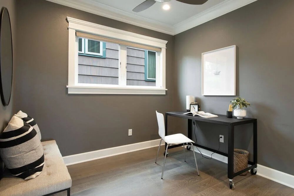 A cozy home office with a gray wall, a window with white trim, a black desk with a white chair, and a small beige cushioned bench with black and white pillows.