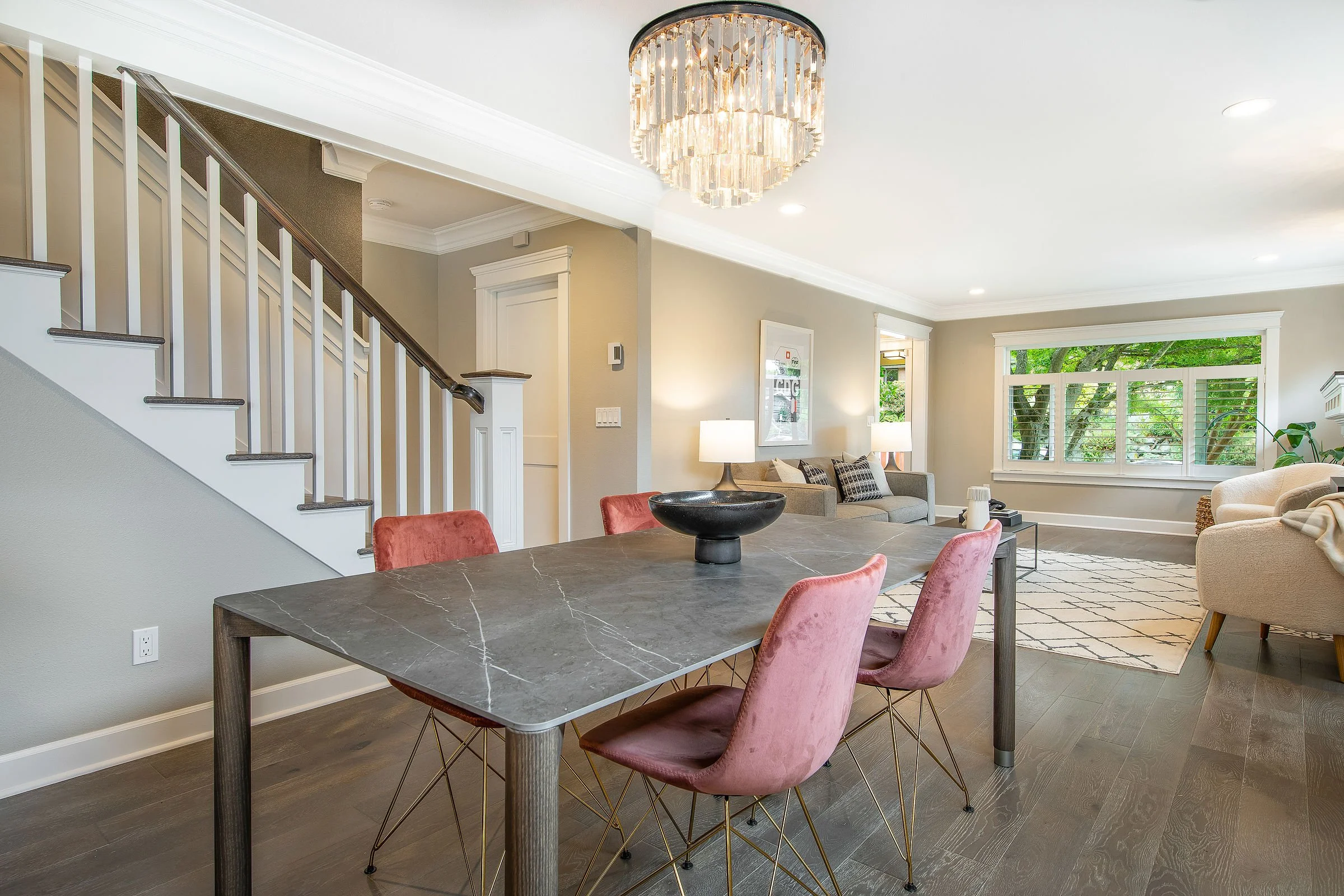 Modern living room with a marble dining table surrounded by pink velvet chairs, a grey sofa with cushions, a large window with white shutters, a chandelier with crystals, and indoor plants, with hardwood floors and light-colored walls.