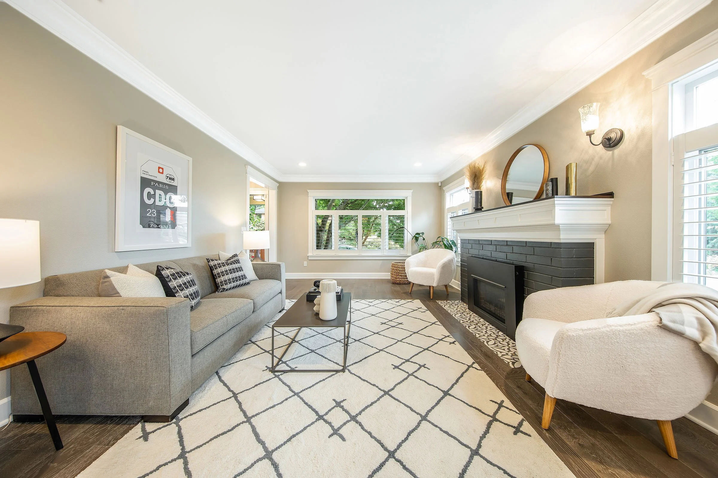 Living room with beige walls, large windows, a gray sofa with patterned pillows, a white armchair, a fireplace with a mirror and decorative items on the mantle, a patterned area rug, and a coffee table.