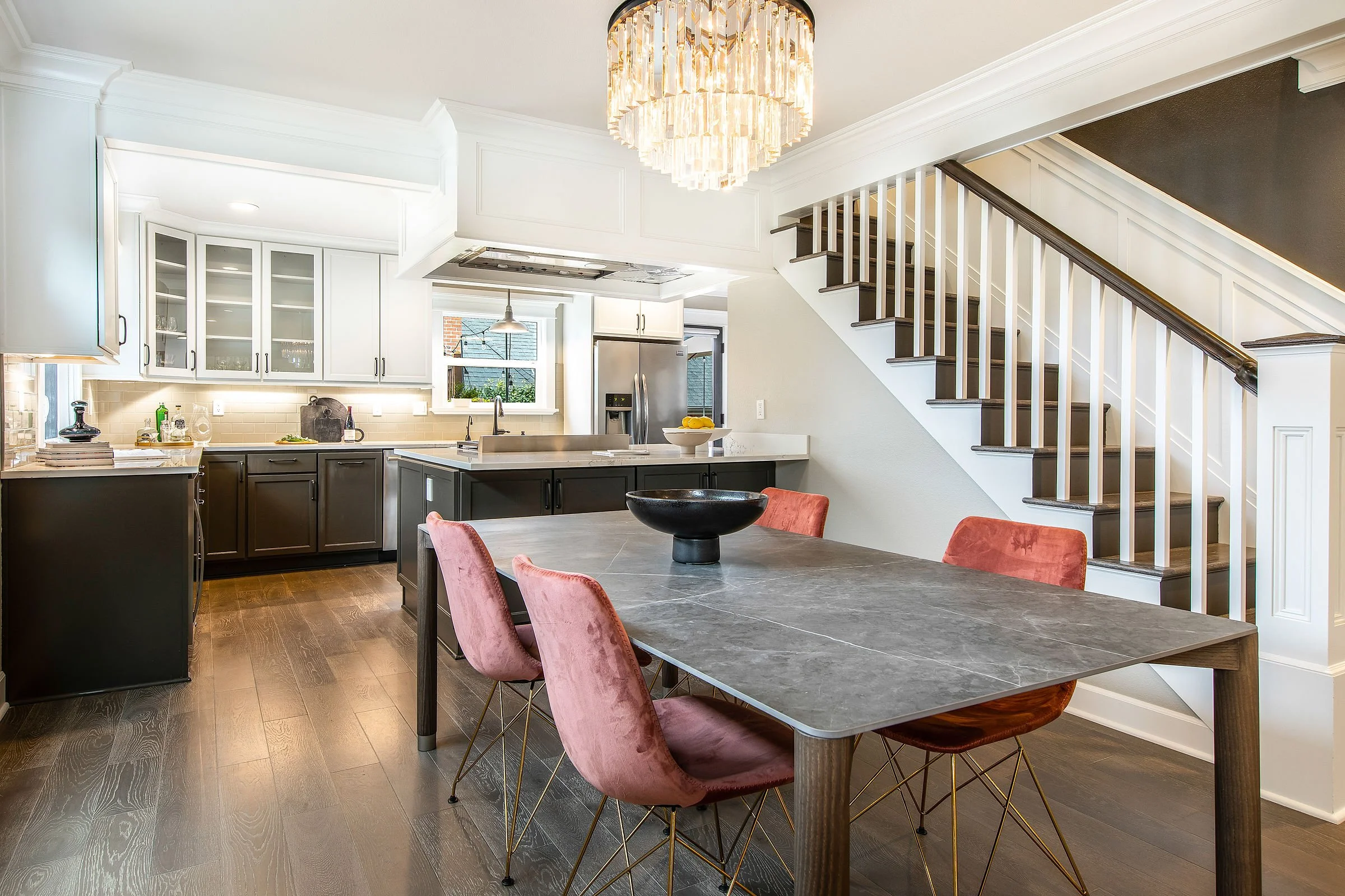 Modern open-concept kitchen and dining area featuring a large gray marble table with pink velvet chairs, white cabinets with glass doors, a stainless steel refrigerator, and a staircase with dark wood steps and white spindles. A large chandelier hang