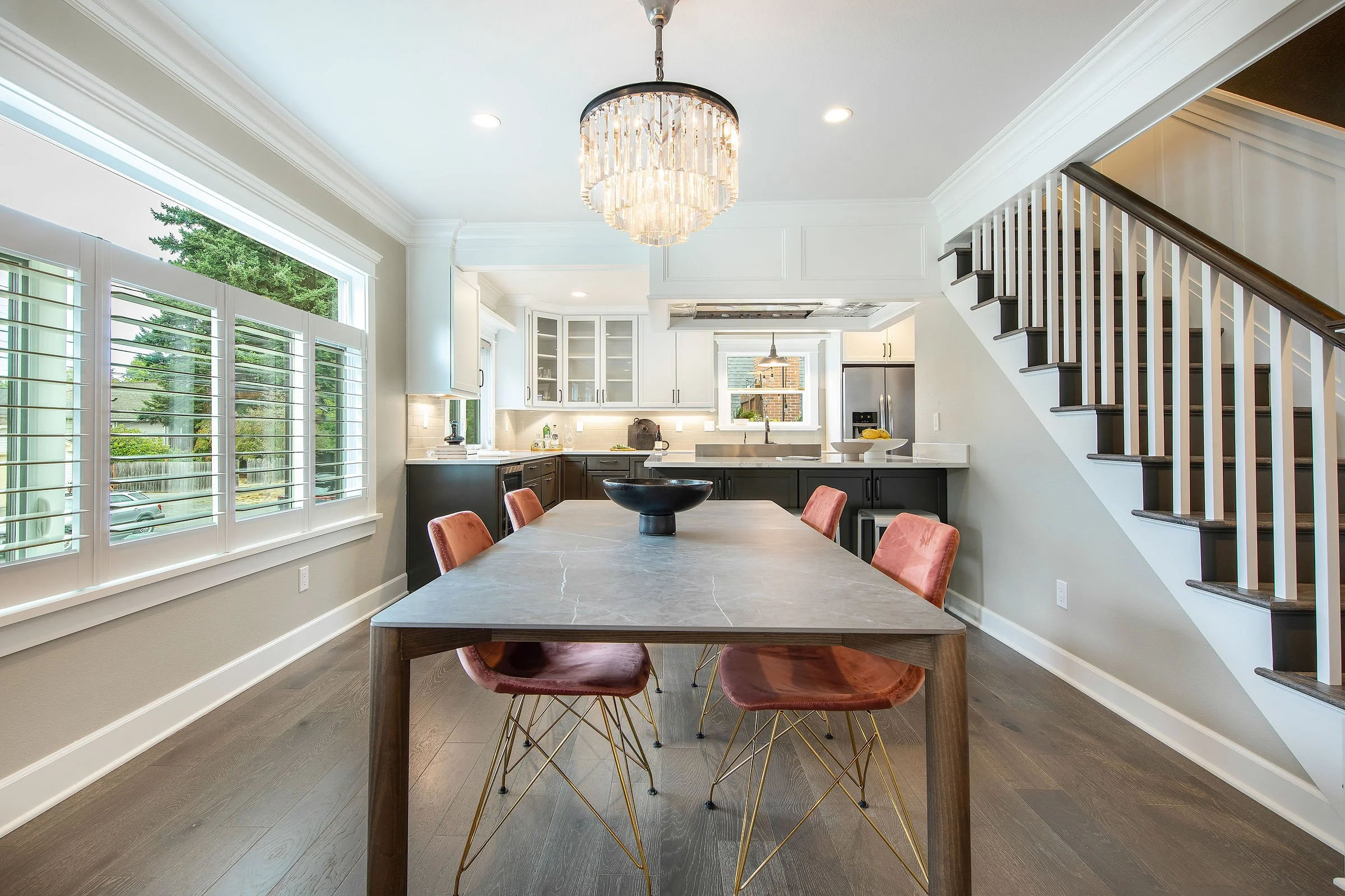 Modern kitchen and dining area with a large rectangular table, pink velvet chairs, white cabinetry, and a staircase, illuminated by natural light from large windows and modern chandeliers.