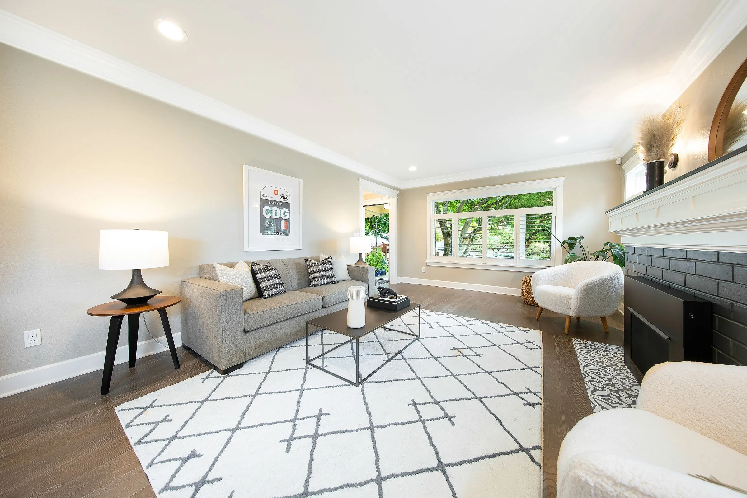 Bright living room with beige walls, large windows with white trim, a gray sofa with patterned pillows, a white armchair, a black fireplace, and a white and black patterned rug.