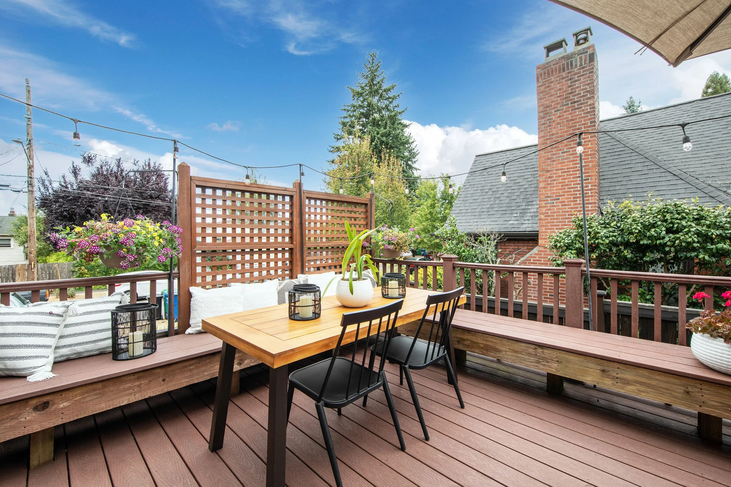 A backyard patio with a wooden dining table, black chairs, potted plants, and string lights under a blue sky with some clouds. There's a brick chimney on the house and a lattice privacy screen with hanging flower baskets.