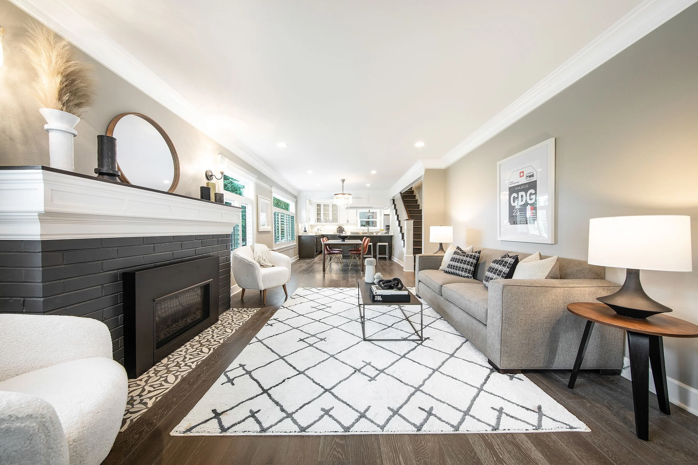 A spacious, modern living room with a black fireplace, white walls, and dark wood flooring. The room features a large light gray sofa with patterned cushions, a white armchair, and a glass coffee table with decorative items. Artworks and a framed pos