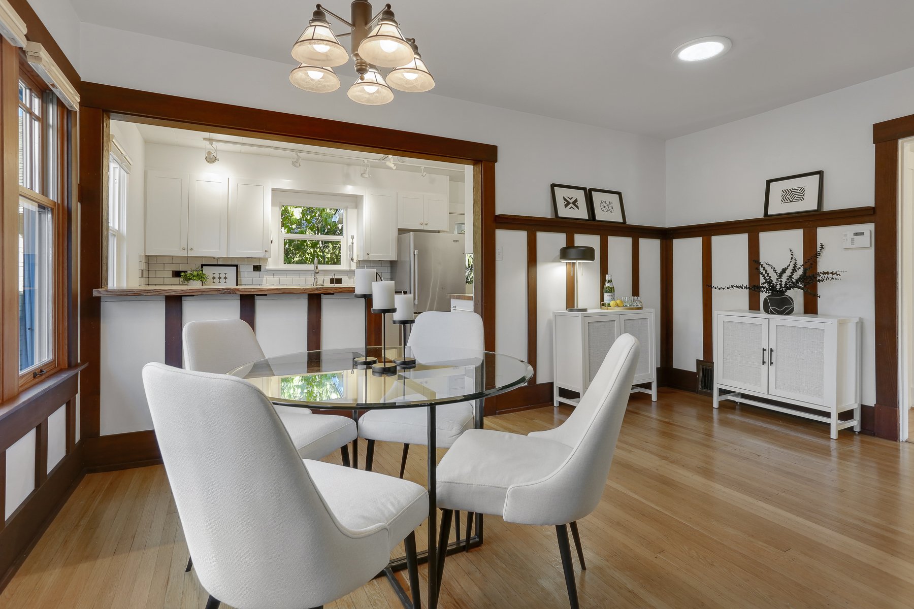 A dining room with a glass-top table and four white upholstered chairs. In the background, a kitchen with white cabinets, a window, and a stainless steel refrigerator. Decor includes black-and-white framed artwork, a black table lamp, and decorative 