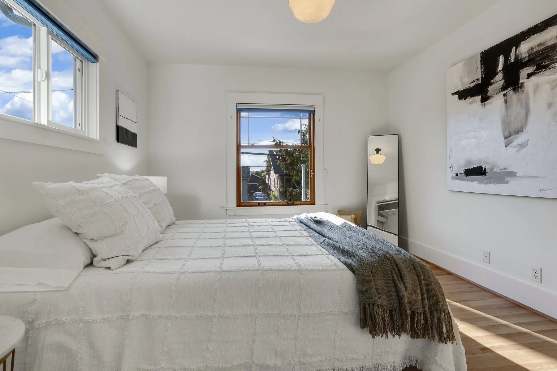 A bright, minimalist bedroom with white walls, a bed with white bedding and pillows, and two windows showing a view of houses and blue sky. Decor includes abstract black and white artwork and a tall mirror.