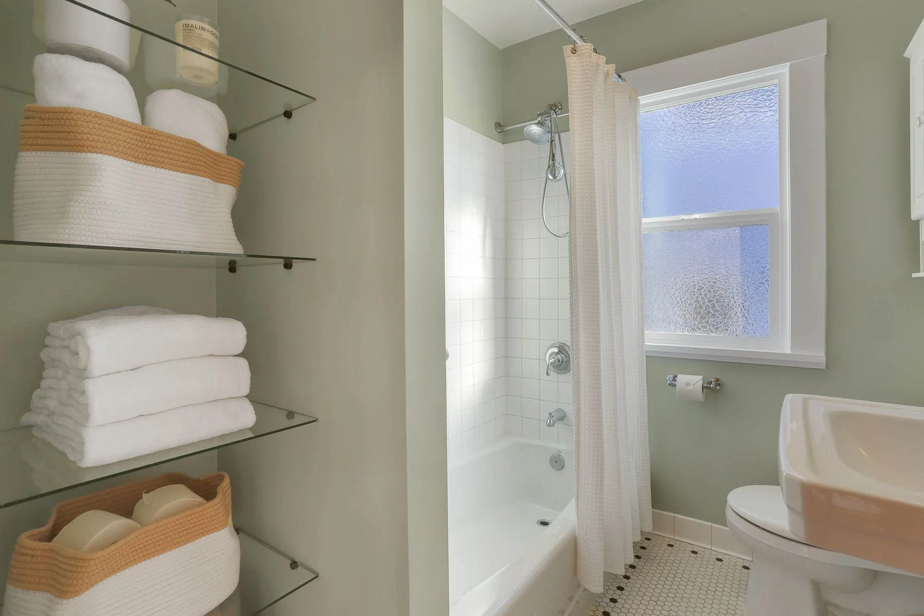A bathroom with white tiles and a bathtub with a showerhead, a window with frosted glass, and a green wall. There are glass shelves on the left with towels and toiletries. A toilet paper roll is on a holder, and a sink is partially visible.
