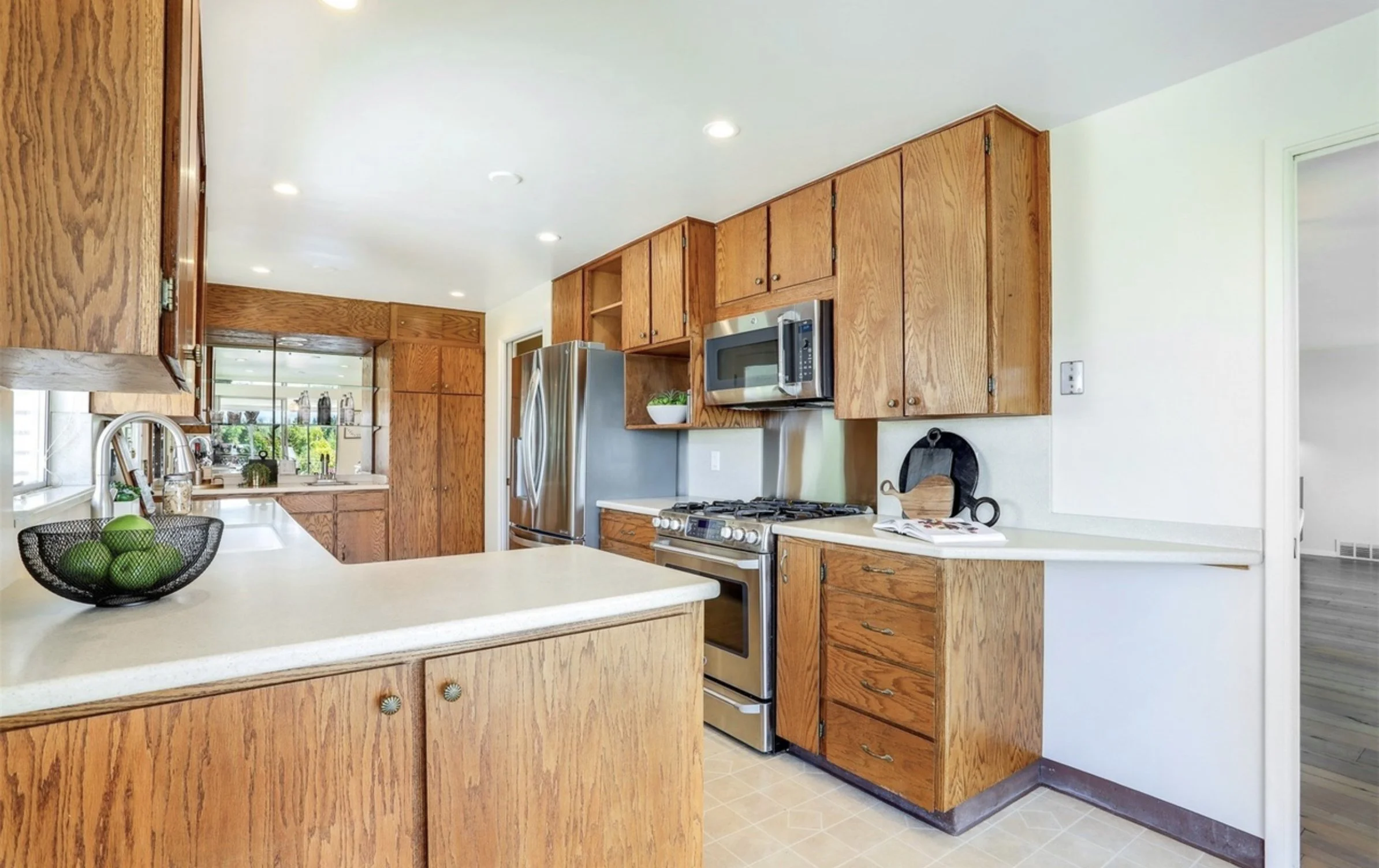 A kitchen with wooden cabinets, a white countertop, a stainless steel refrigerator, microwave, gas stove, a bowl of green apples, and a window with a view of a green yard.