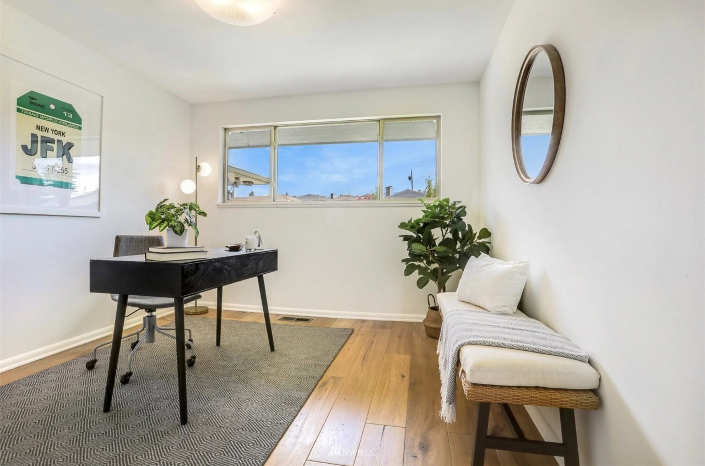 A cozy living room with white walls and hardwood flooring, featuring a black desk with books and a plant, a gray armchair, a white daybed with pillows and a blanket, a round mirror on the wall, a framed poster, large window with a view of a neighbori