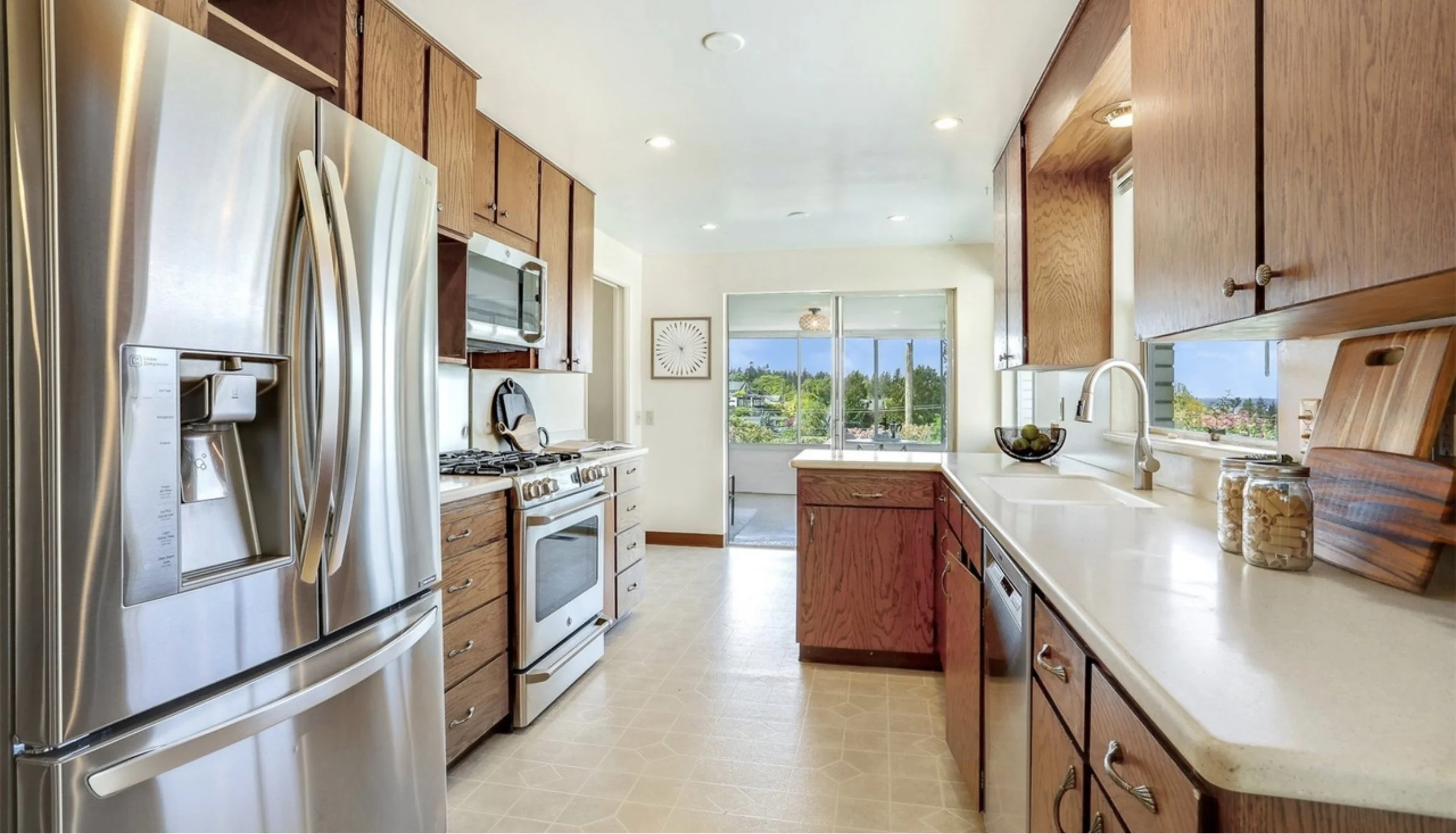 A kitchen with wooden cabinets, stainless steel refrigerator, stove, microwave, and white countertops. Large windows provide a view of greenery outside.
