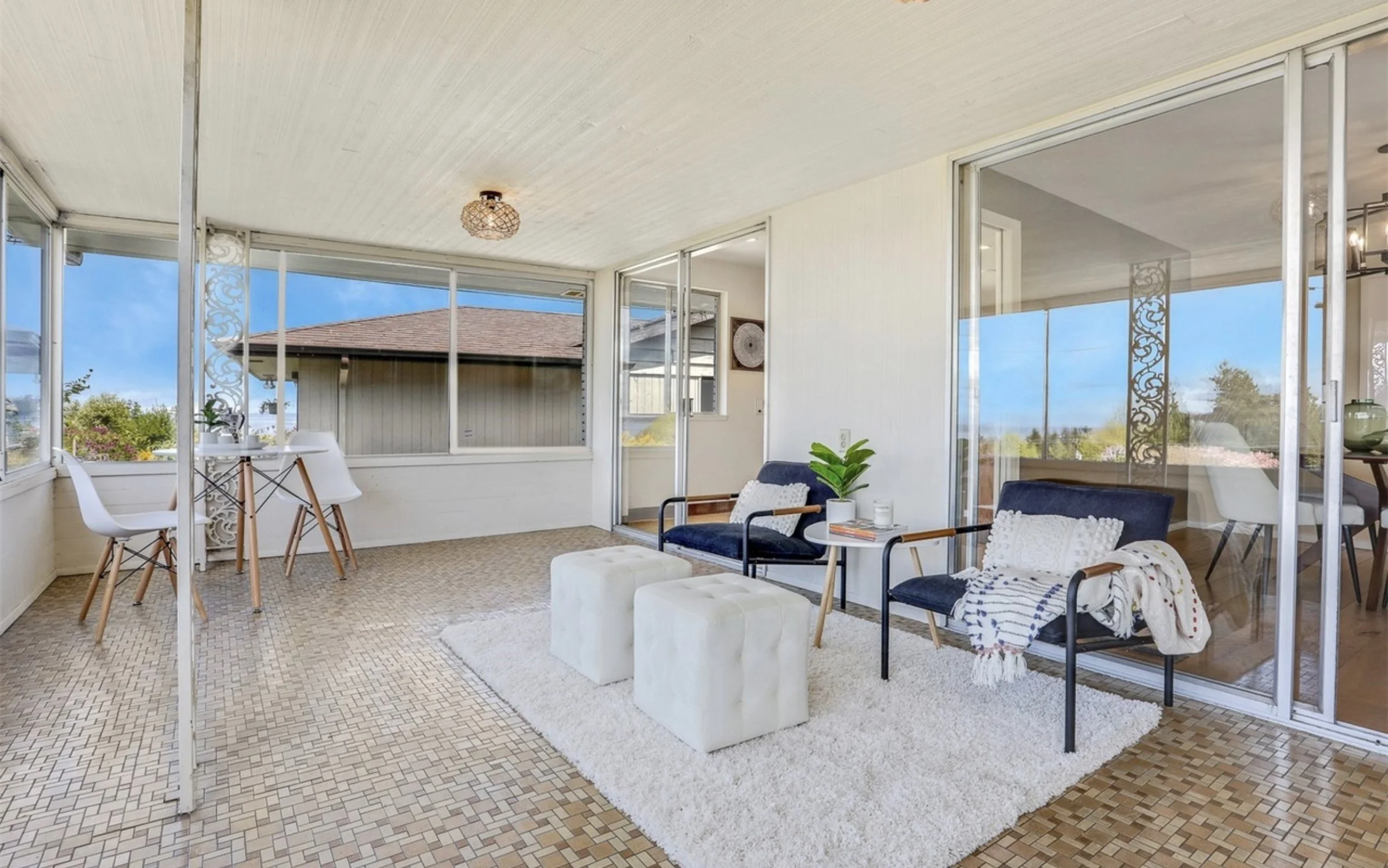 Bright enclosed porch with large windows, wicker ceiling light, sitting area with dark blue chairs, white ottomans, white side table with books, houseplants, and outdoor view.