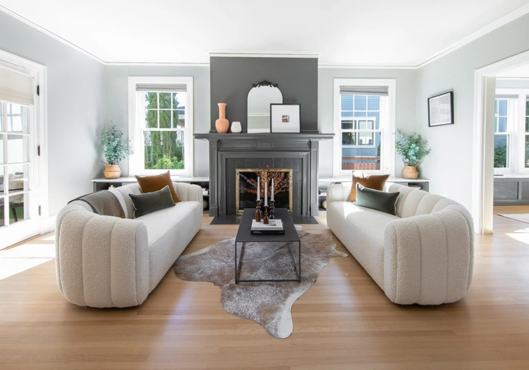 Living room with two white sofas facing each other, a black coffee table in the center, a cowhide rug, a gray fireplace with decorative items, and large windows with greenery outside.