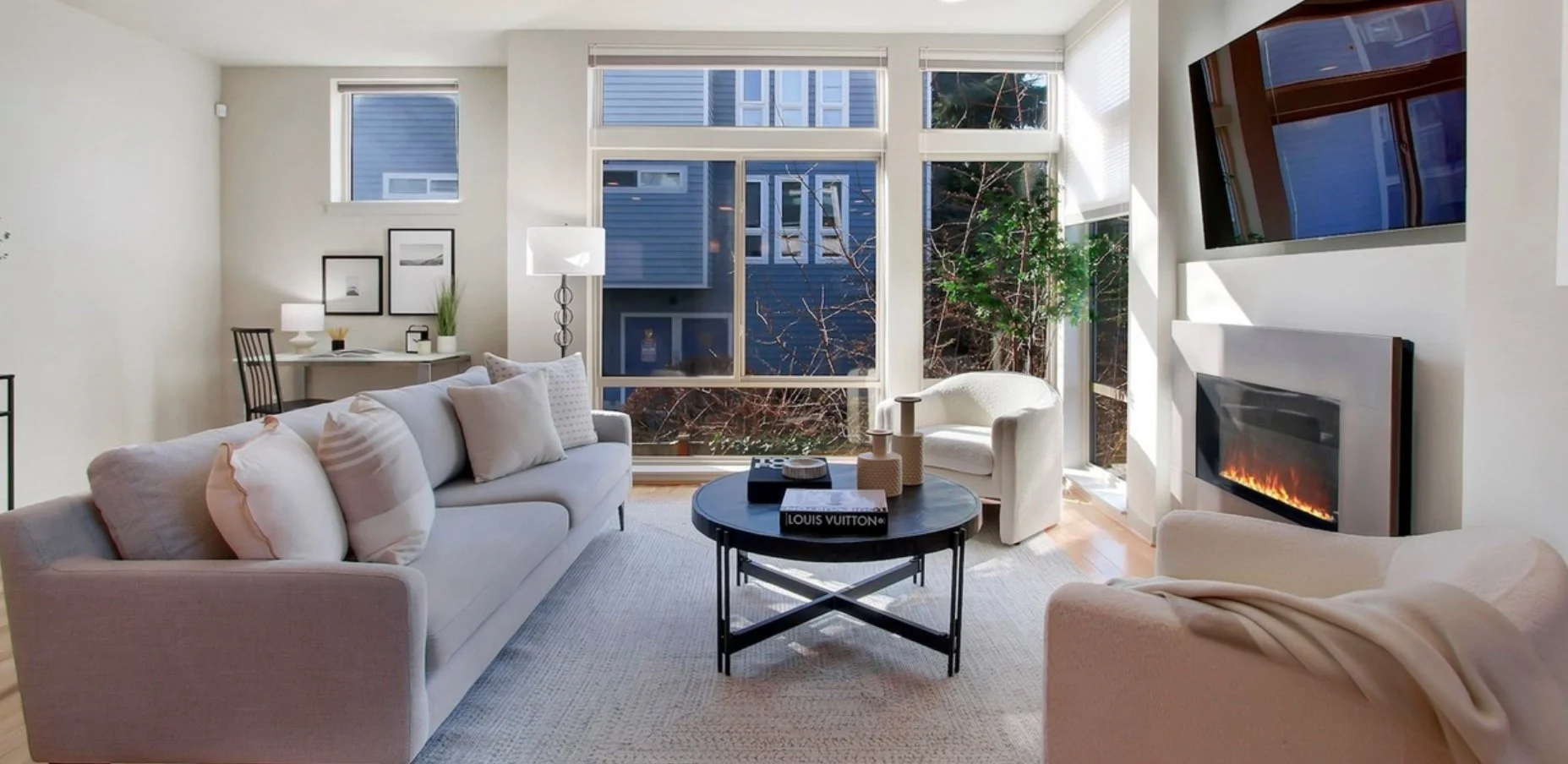 Bright living room with large window, white sofa and cozy chairs, a black coffee table with decor, and a wall-mounted TV and fireplace.