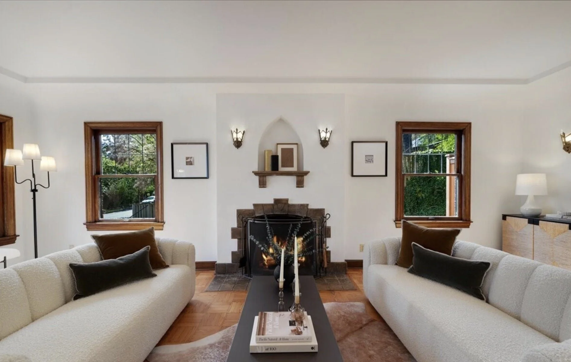 Living room with two white sofas, a fireplace with a fire, two windows with wooden frames, and cozy decor including framed art, lamps, and books.