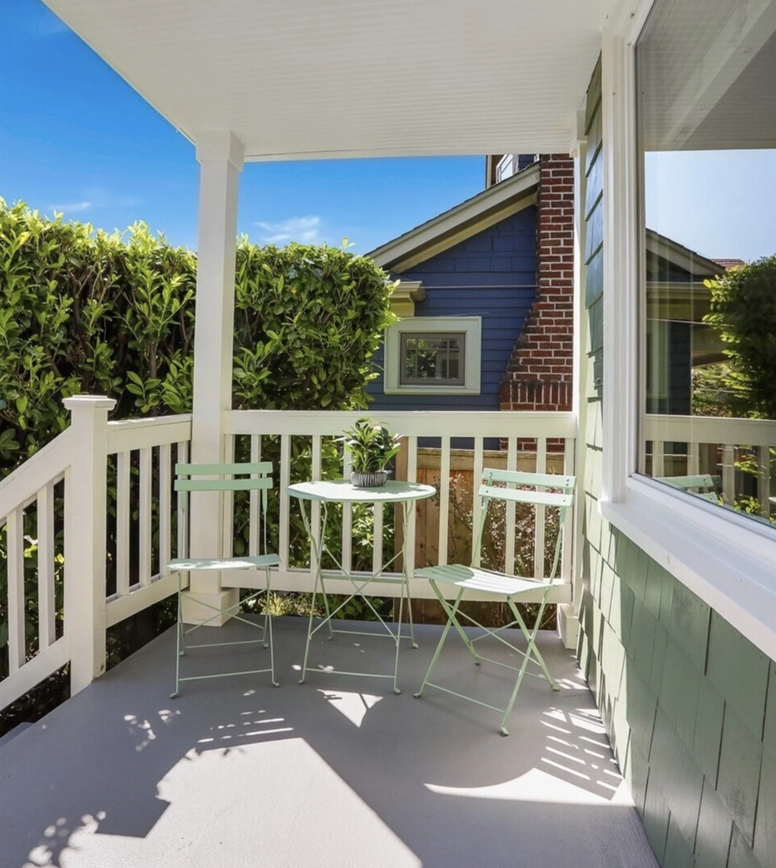 A small outdoor balcony with white railing, a round white table, and three light green chairs. A potted plant sits on the table. The balcony overlooks a garden with bushes and neighboring houses, under a clear blue sky.