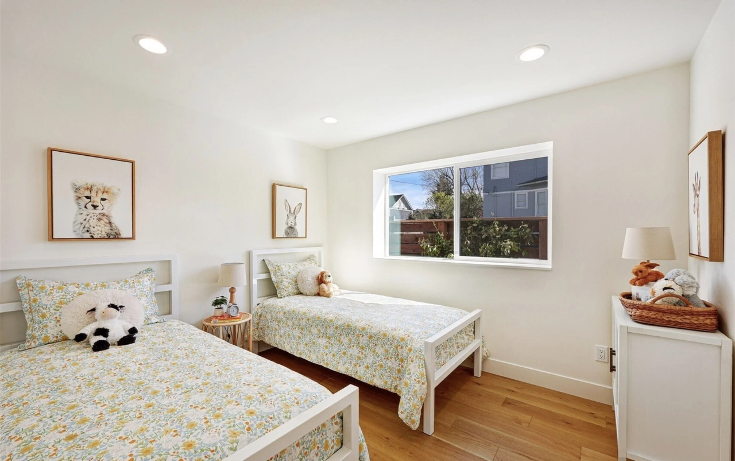 A bright children's bedroom with two twin beds covered in floral bedding, each with stuffed animal toys and pillows, decorated with animal-themed framed art on the walls, a window with a view of a house and trees, and a wooden floor.