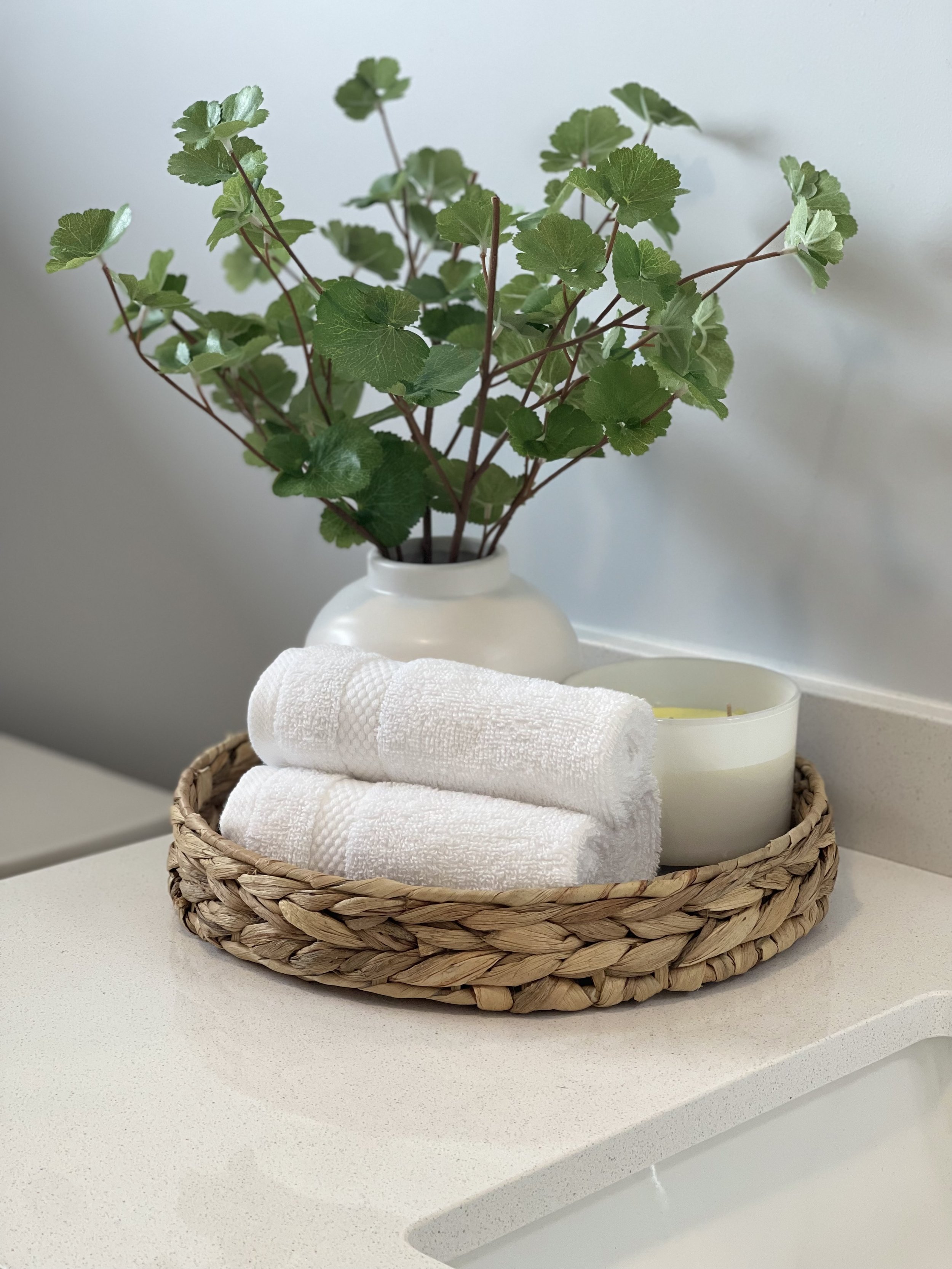 A wicker basket on a white countertop holding three white folded towels, a small white candle, and a potted plant with green leaves in a white vase.