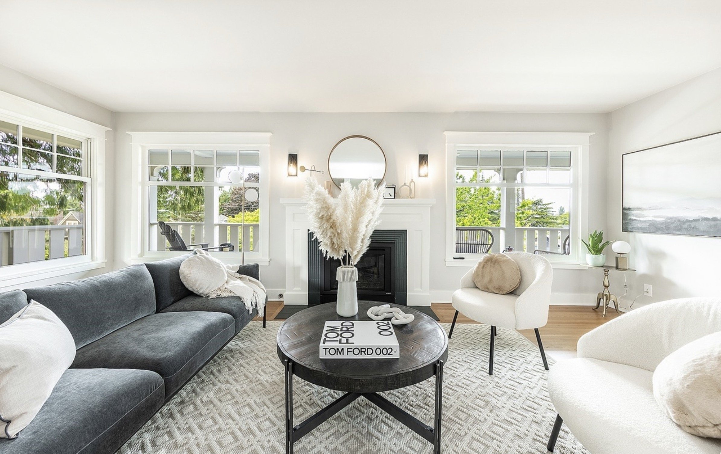 Bright living room with large windows, white walls, a white fireplace, and modern furniture including a gray sofa and white chairs. Decor includes a round mirror above the fireplace, a vase with pampas grass on the coffee table, and artwork on the wa