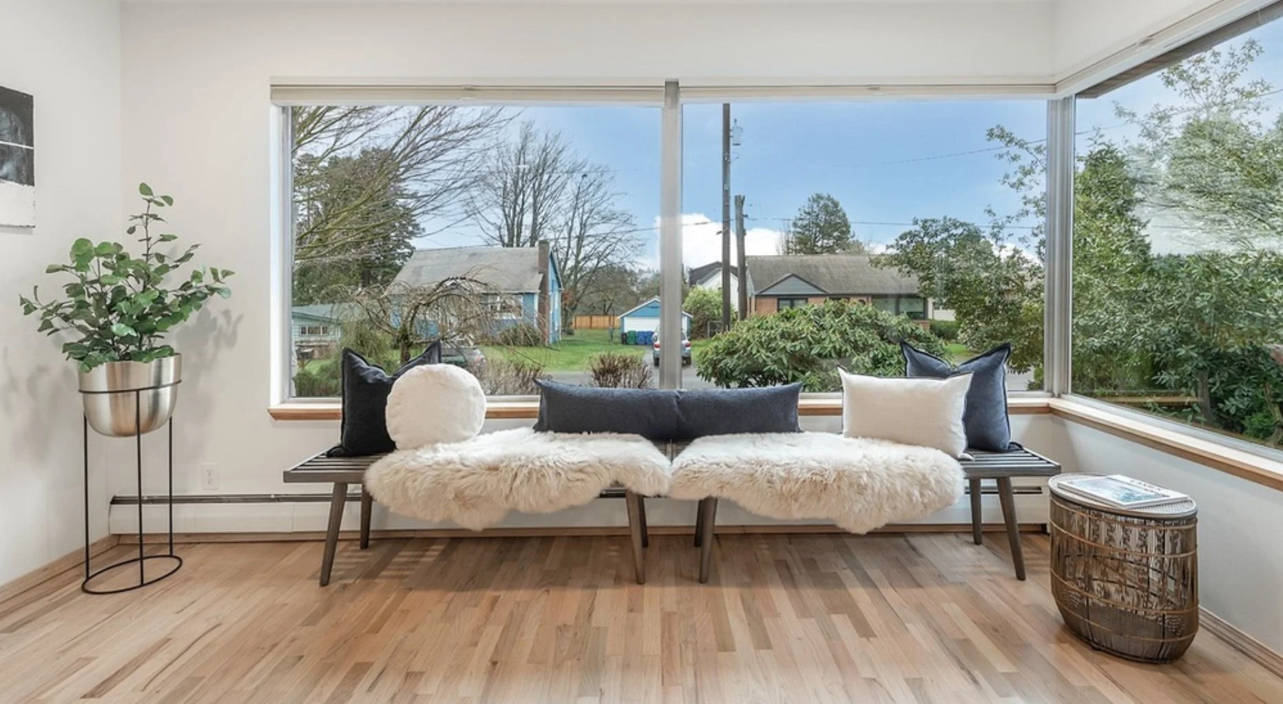 Living room with a large window view of suburban neighborhood, a wooden bench with pillows and fluffy throws, a potted plant on a stand, and a small side table.