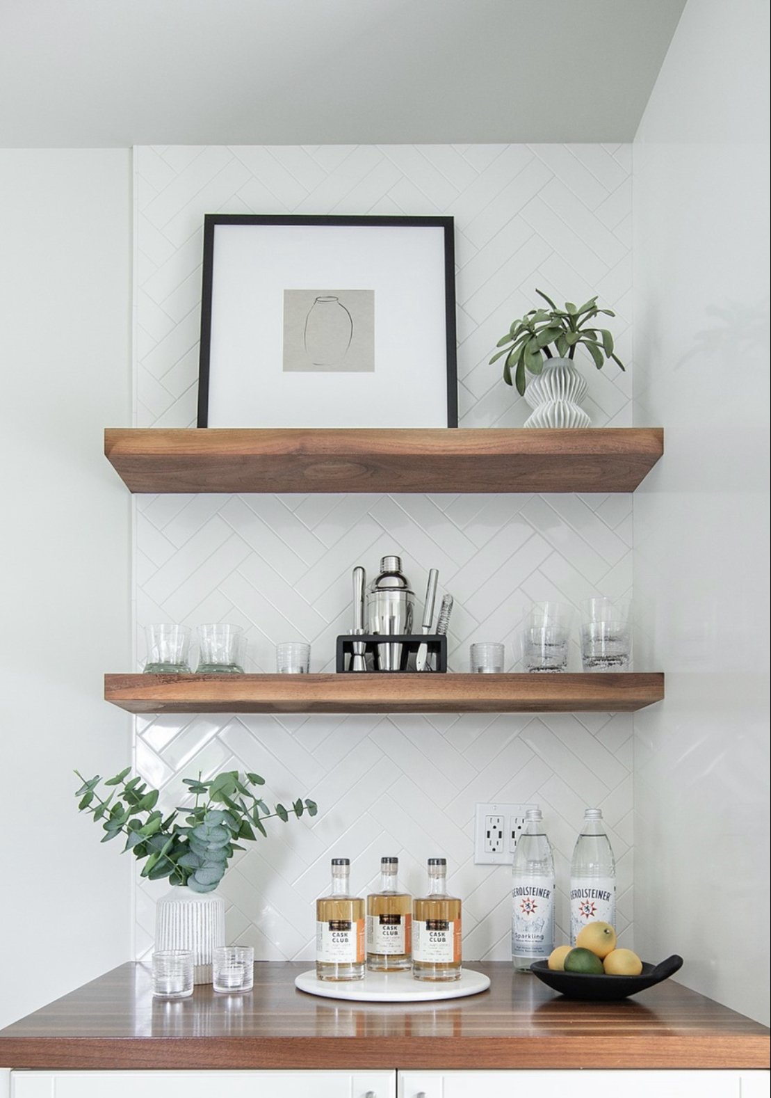 Living room or kitchen wall with two wooden shelves holding a framed sketch of a vase and a decorative green plant in a white vase. Below the shelves are drinks, glasses, water bottles, and a fruit bowl with lemons and limes on a wooden countertop.
