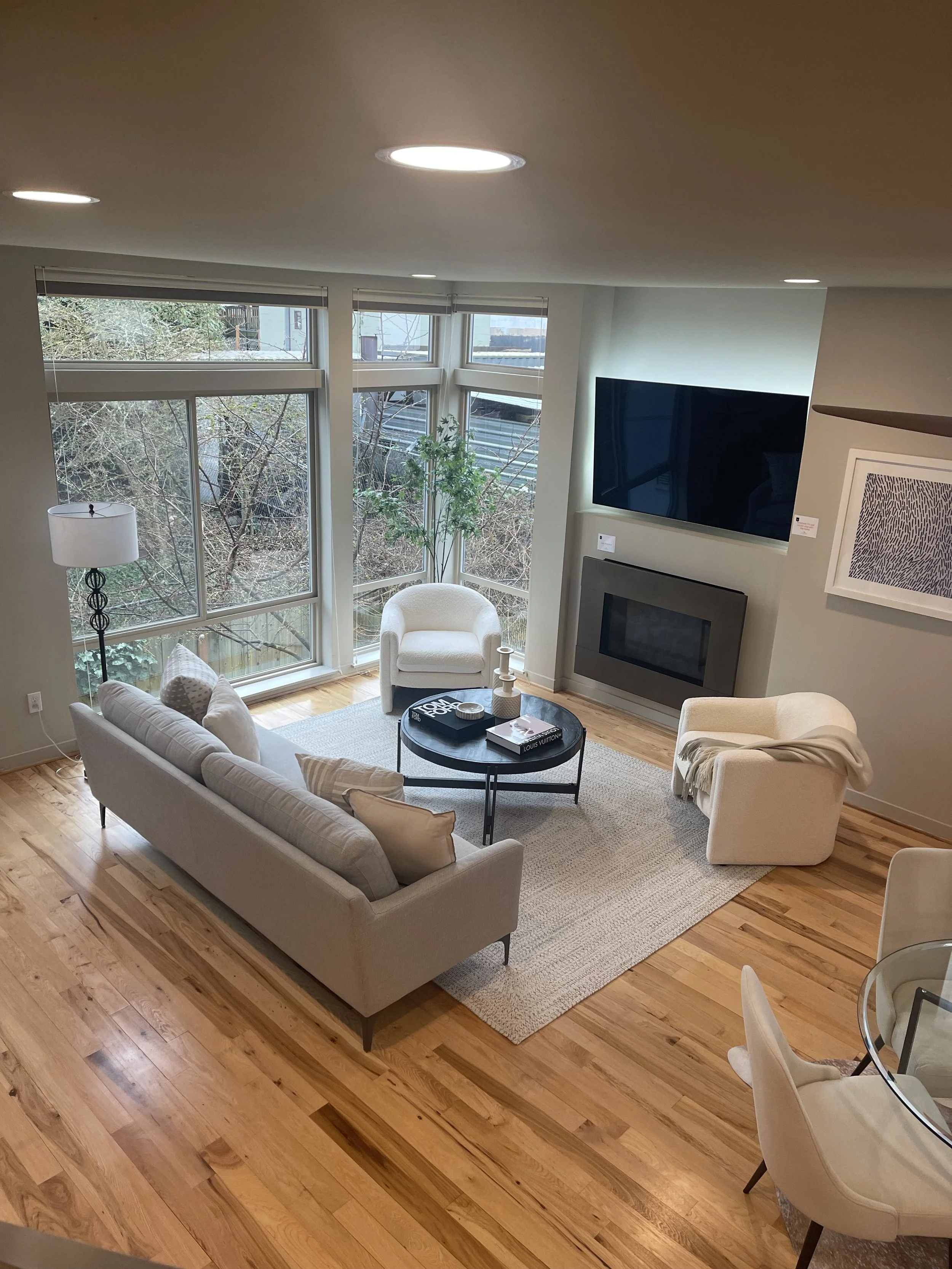 Living room with beige sofa, white armchair, round black coffee table with books and decor, large windows with blinds, wooden floor, fireplace, mounted flat-screen TV, tall floor lamp, abstract framed artwork, part of dining area with white chairs.