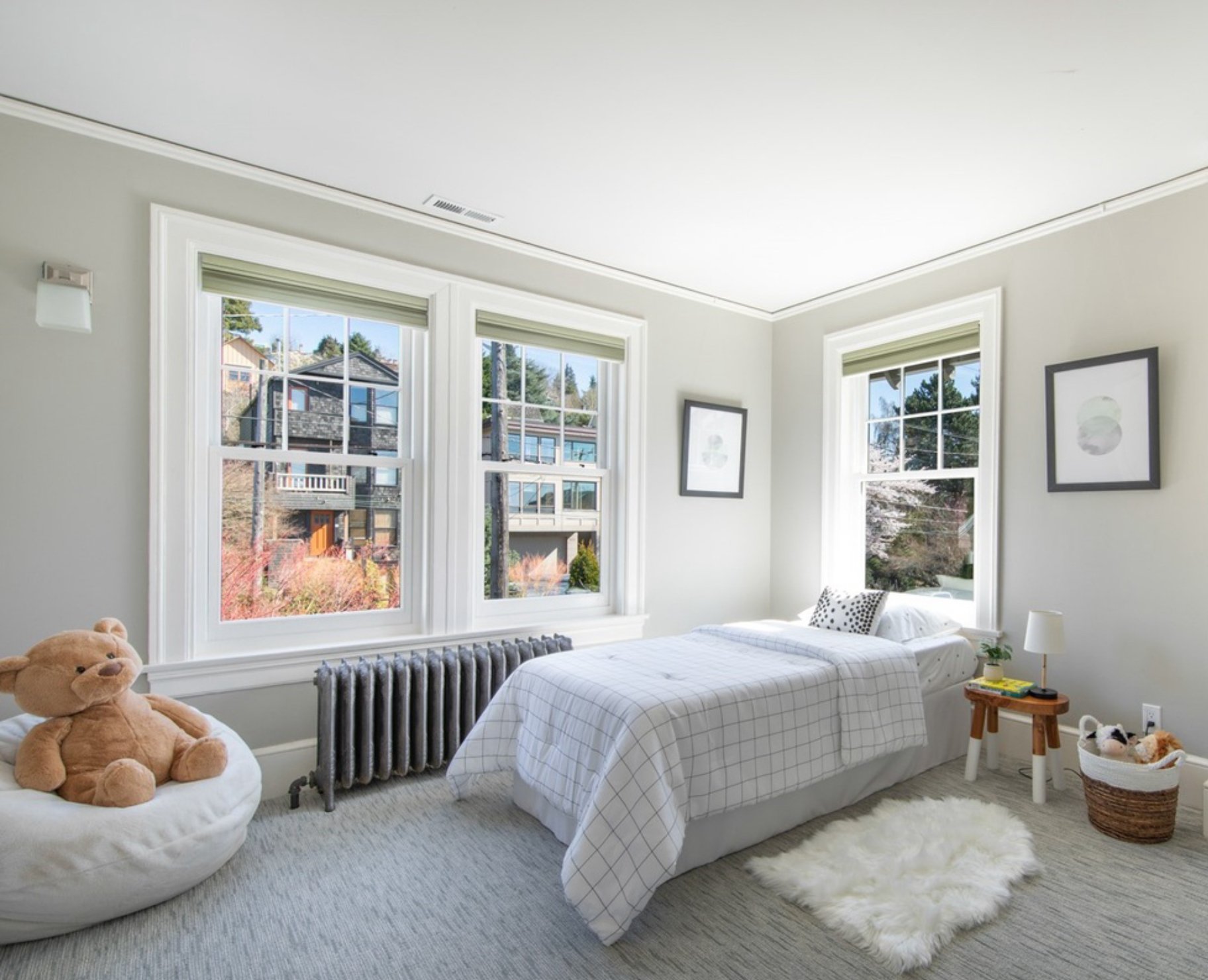 A cozy bedroom with three large windows showing a neighborhood view, a bed with a checkered white and gray bedspread, a stuffed bear on a round white chair, framed art on the walls, a small side table with a lamp and books, a shaggy white rug, and a 