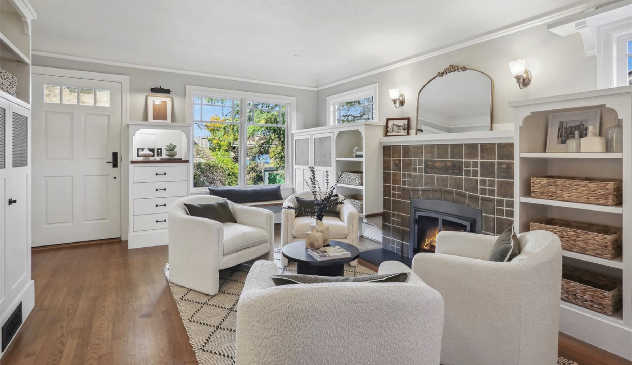 Living room with white sofas around a black coffee table, wood flooring, large windows showing greenery outside, a brick fireplace, built-in white shelves with decorative items, and a mirror above the fireplace.