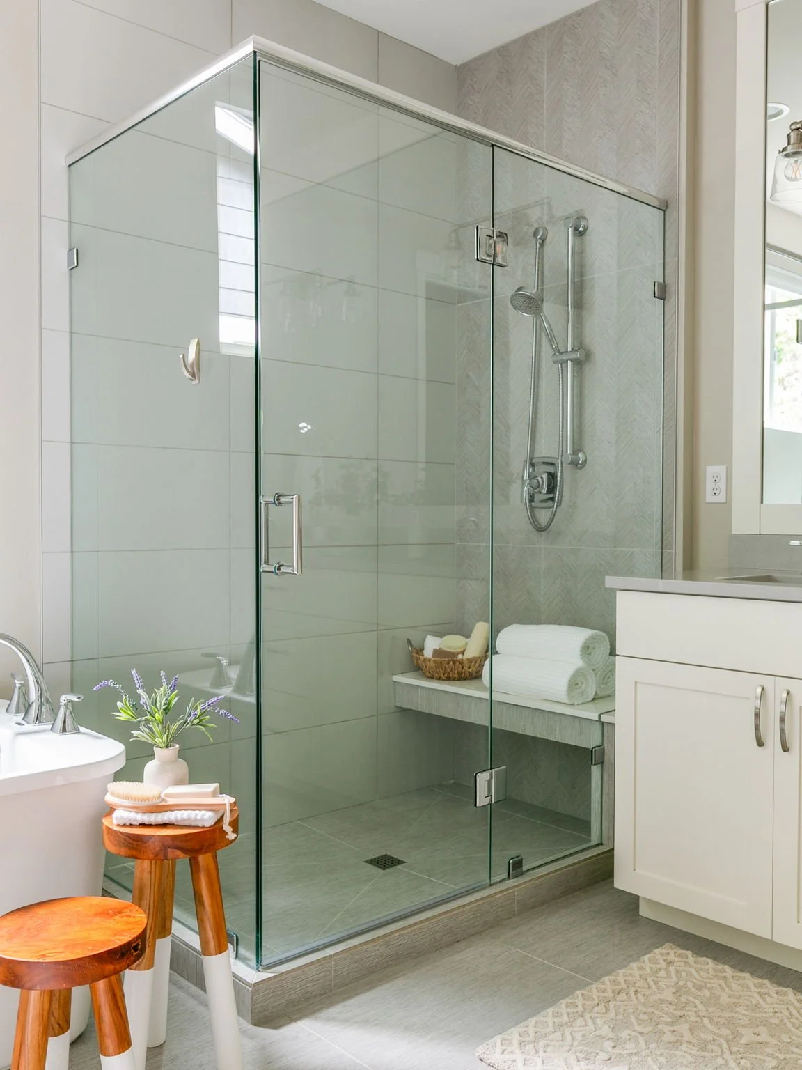 Modern glass-enclosed shower with a rain shower head and hand-held shower, white towels in a basket, and potted plant on small wooden stools in a clean, bright bathroom.