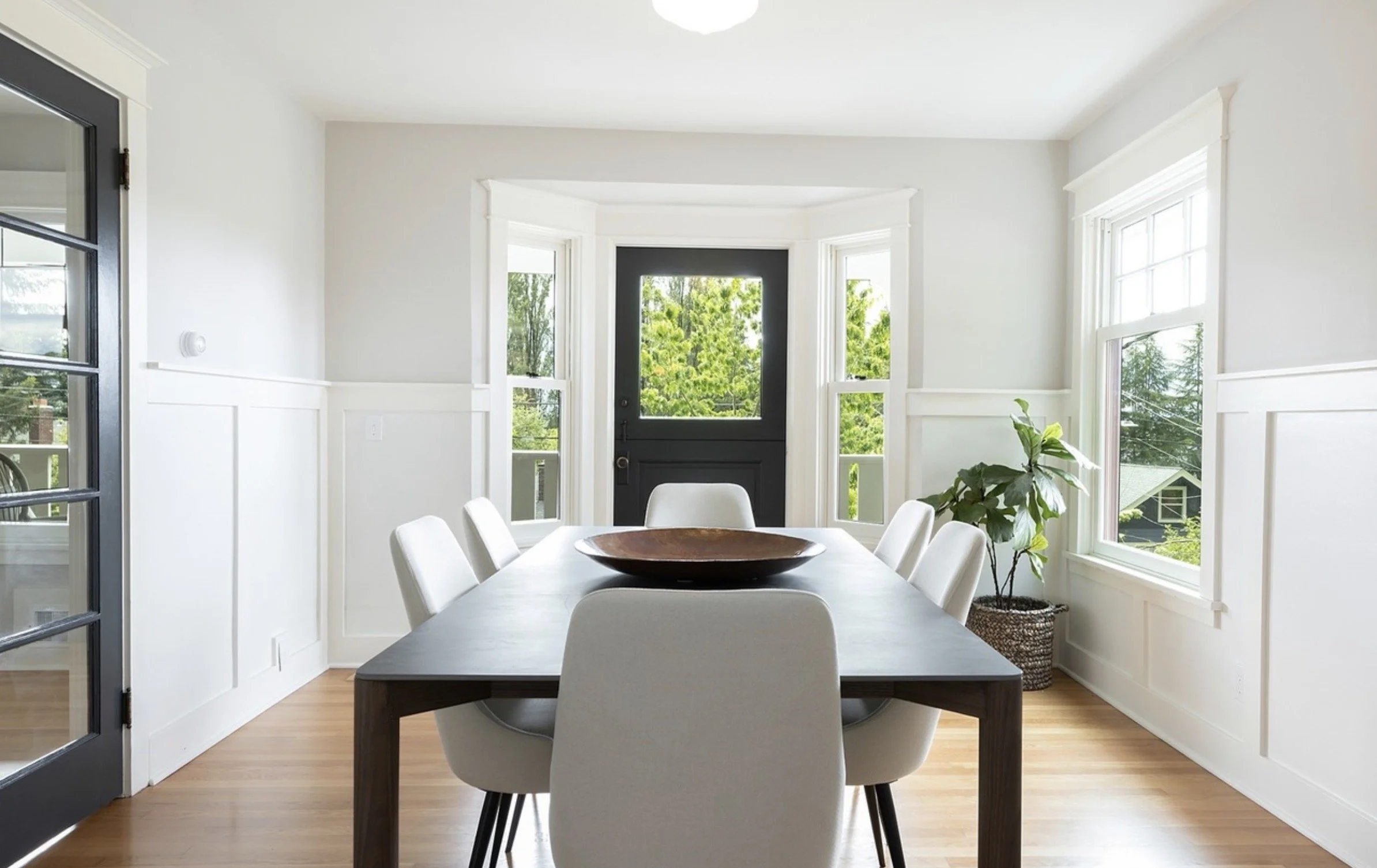 A dining room with white walls, large windows, a black door, and a rectangular dining table with white chairs. A potted plant is near the window.