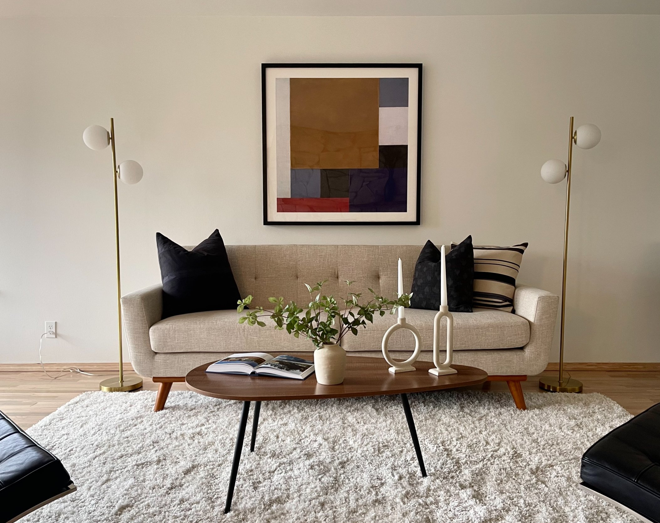 Living room with a beige sofa, black pillows, a wooden coffee table with white vases and an open book, two tall gold-and-white floor lamps, a large abstract framed art piece on the wall, a white textured rug, and hardwood floors.