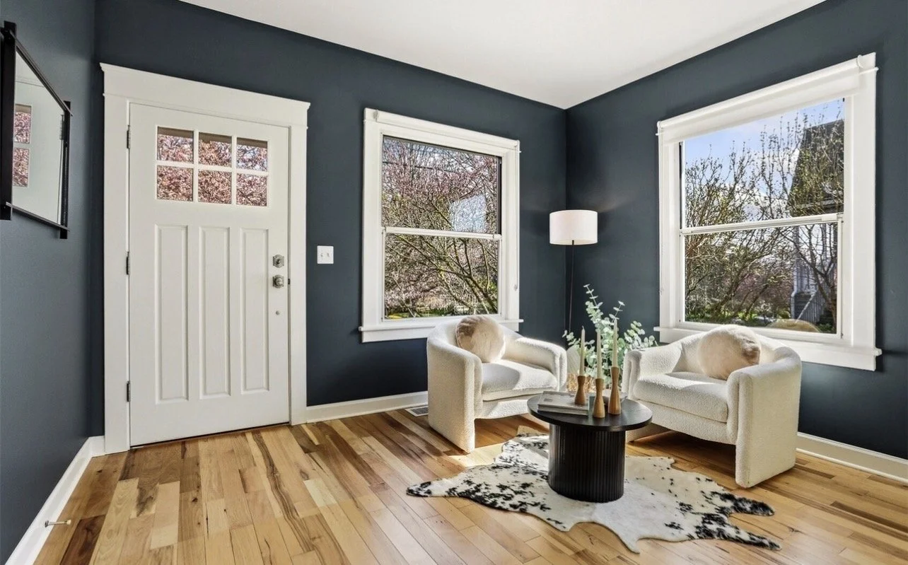 Living room with dark blue walls, two white armchairs, a cowhide rug, a black round coffee table with vases, a tall white floor lamp, and large windows showing trees outside.