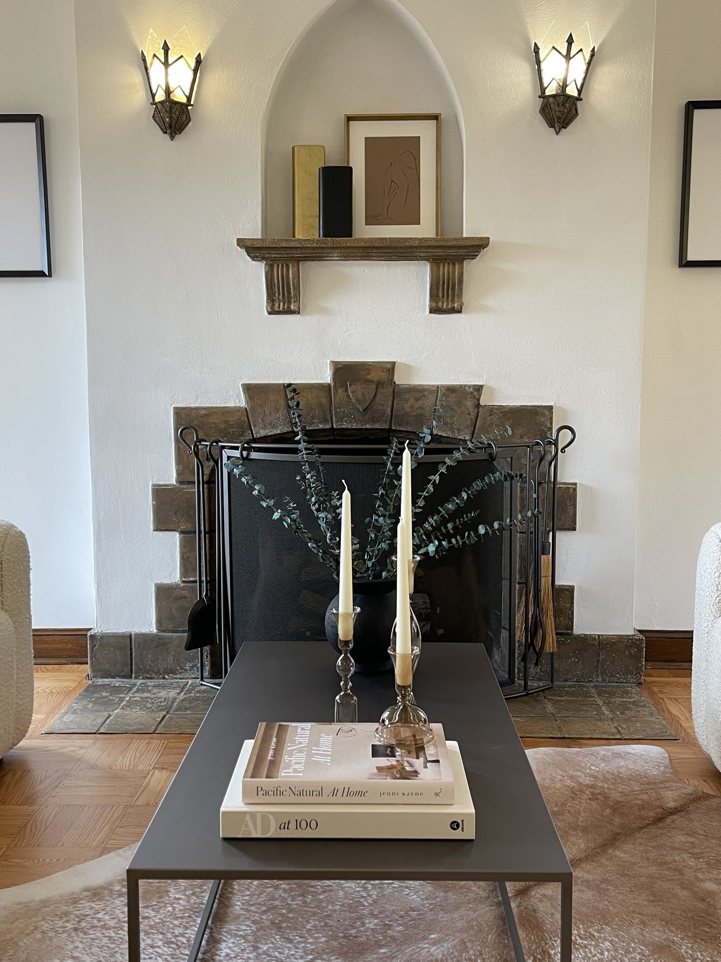 A living room with a fireplace, a black table with two books, a black vase with green leaves, and two white candles. The fireplace has a beige brick surround and a black screen. Above the mantel, there are two black and gold vases, a framed artwork, 