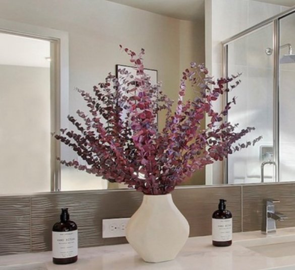 Vase with purple and pink artificial flowers on bathroom countertop with mirror, soap dispenser, and shower in background.