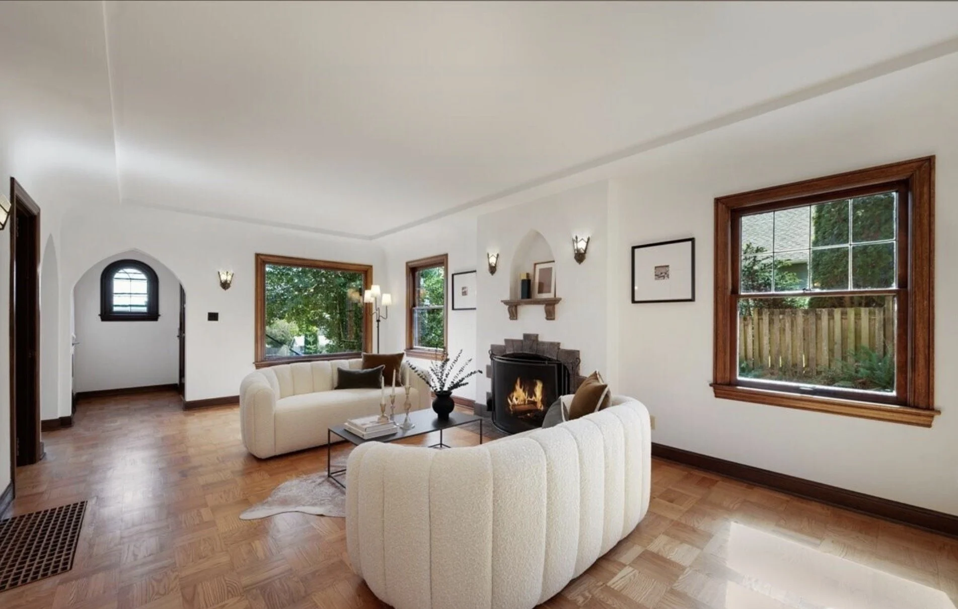 A living room with white walls, large wooden-framed windows, a fireplace with a fire burning, and cream-colored sofas arranged around a black coffee table with decorative items, hardwood floors, and some wall art.