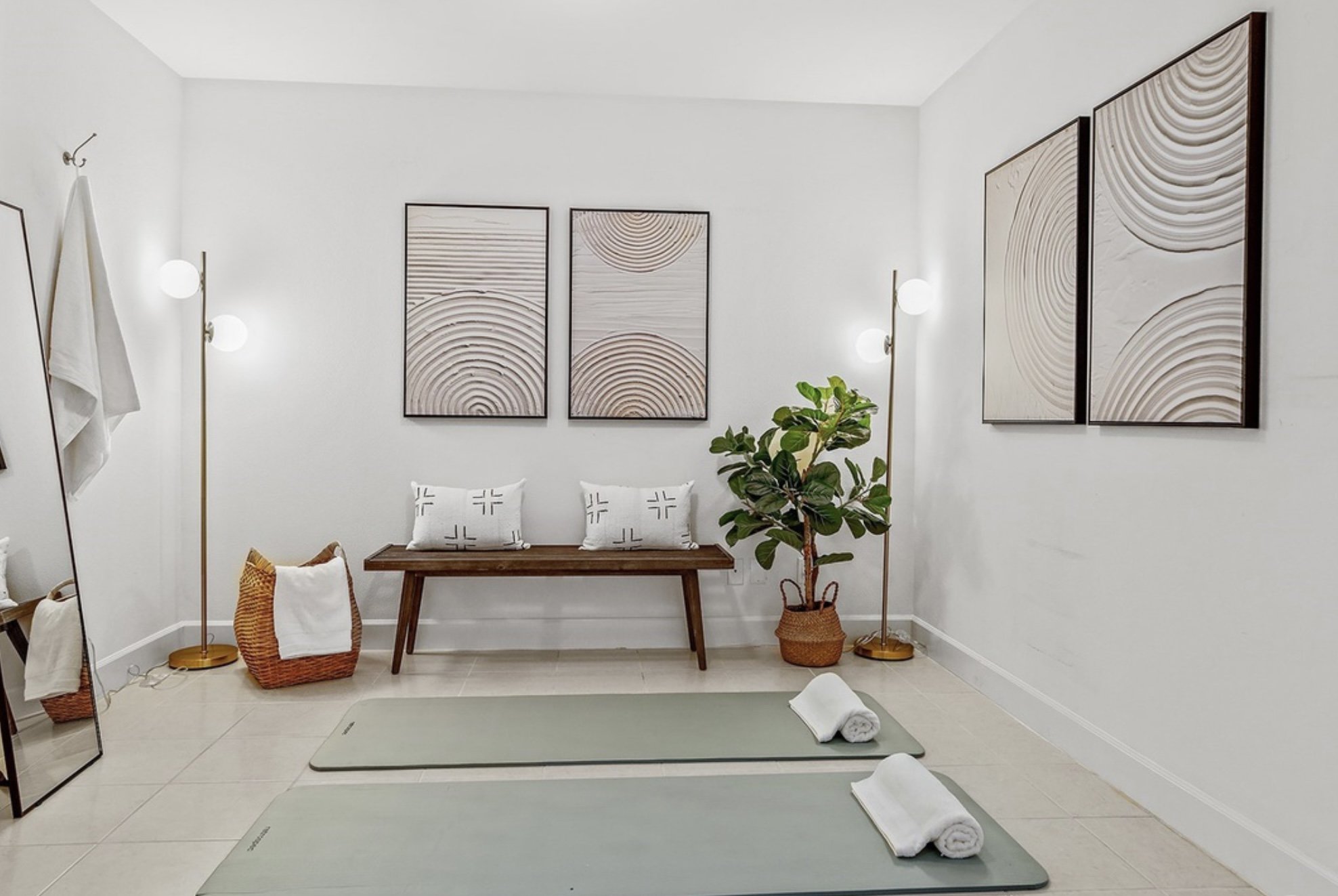 The image shows a minimalist yoga or meditation space with light green mats, rolled towels, and a stylish interior decor. It features framed wall art with abstract circle patterns, a wooden bench with pillows, potted plants, and modern floor lamps wi